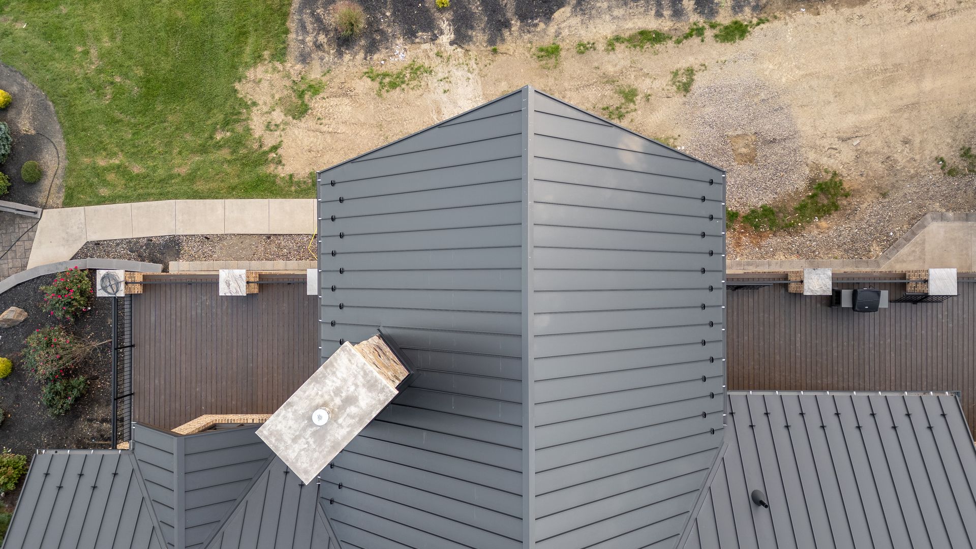 Aerial view of a gray metal roof with a chimney, surrounded by brown decking and a grassy area.
