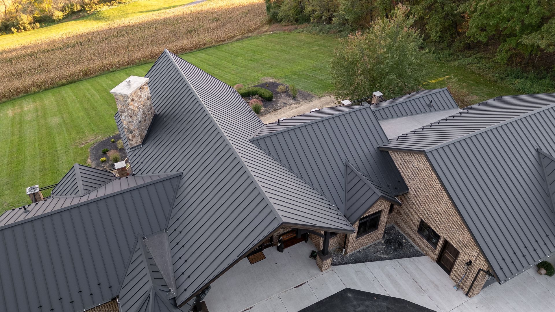Drone shot of a house with a dark gray metal roof and stone chimney, set in a rural landscape.