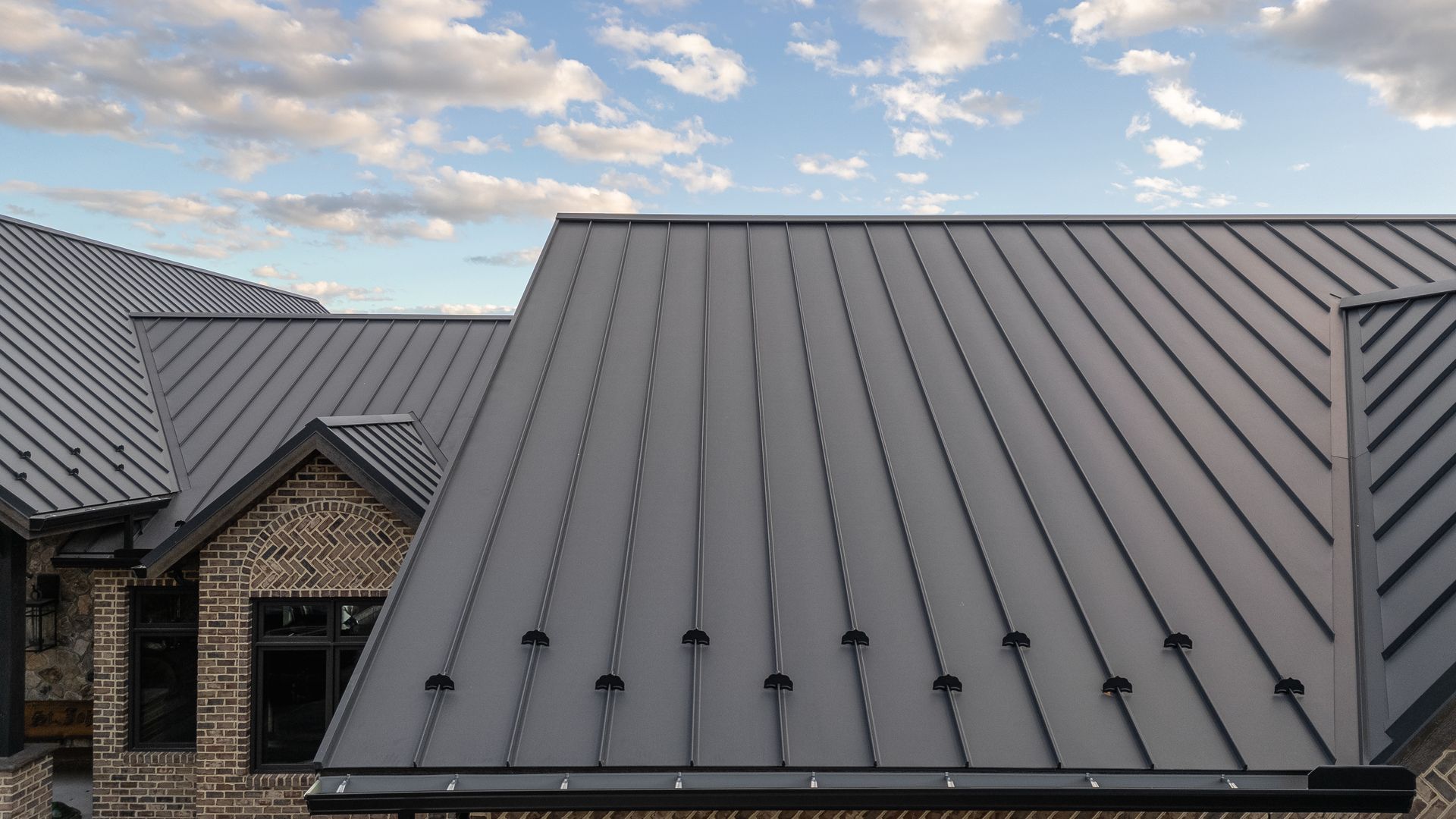 Gray metal roof on a house with brick, against a blue sky with clouds.