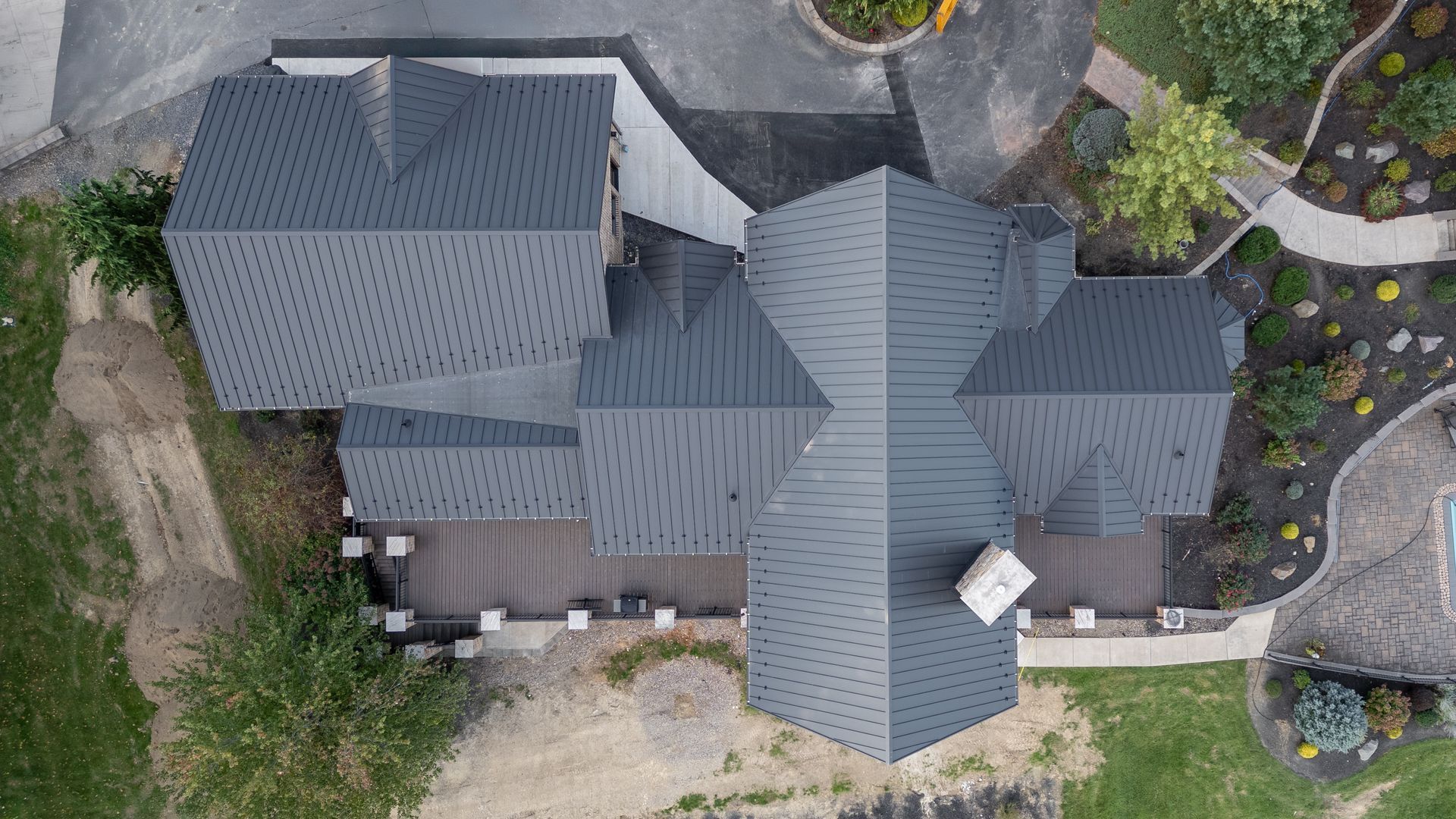 Overhead view of a house with a complex dark gray roof, deck, and landscaping; surrounded by trees and grass.
