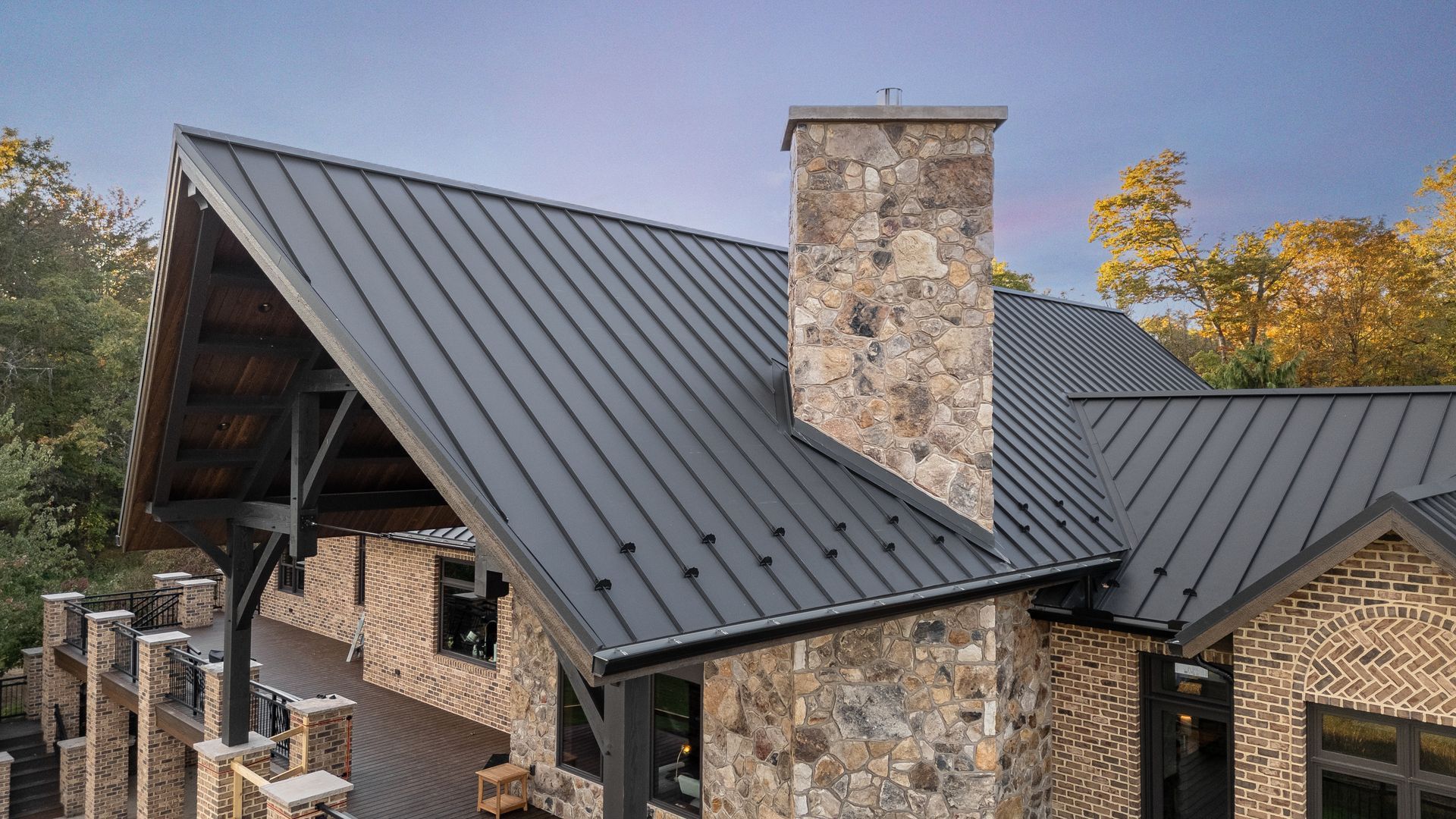A modern house with a black metal roof, stone chimney, and brick exterior, set against a tree-filled background.