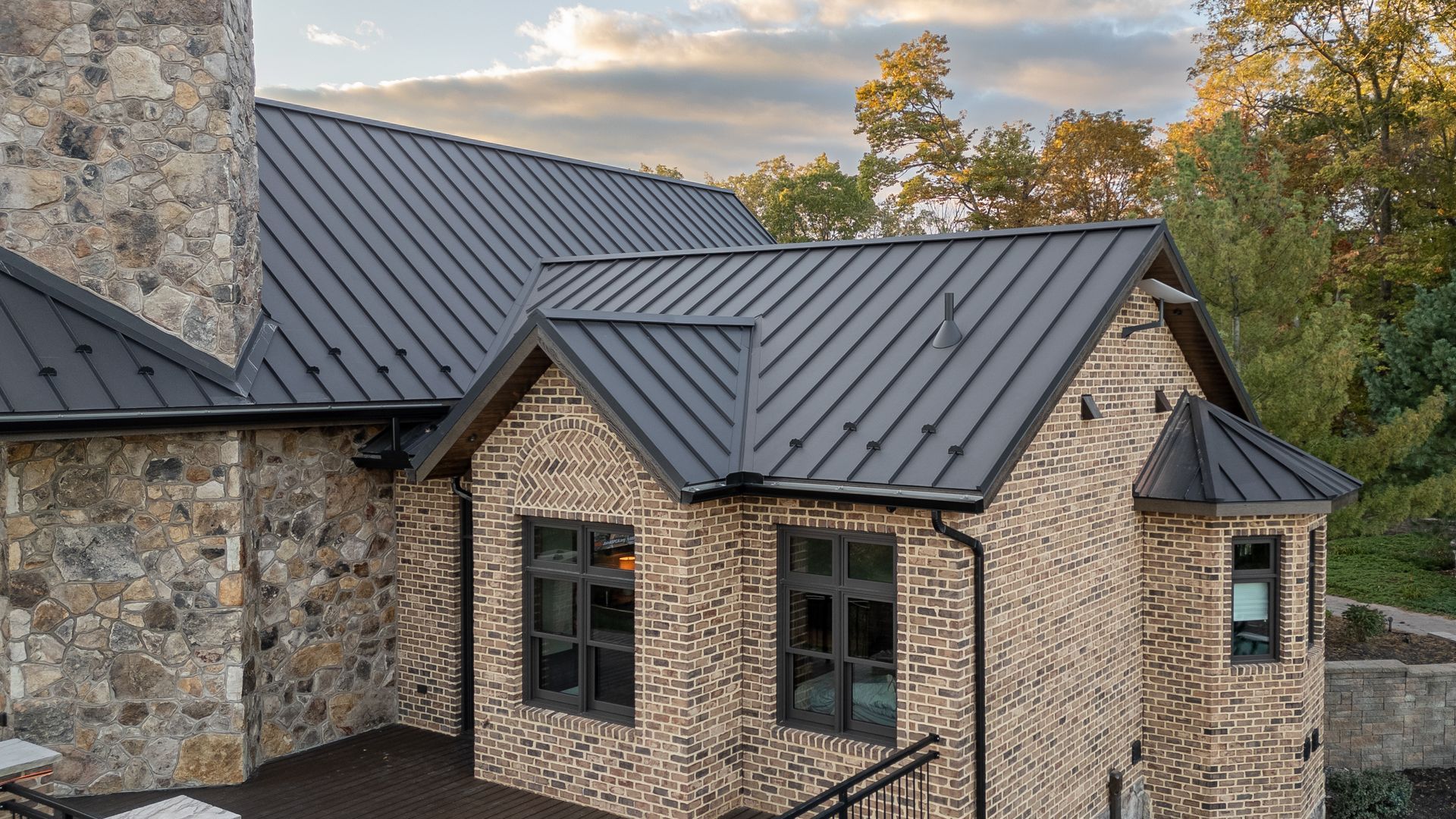 Brick house with dark gray metal roof, windows, and stone chimney under a cloudy sky.