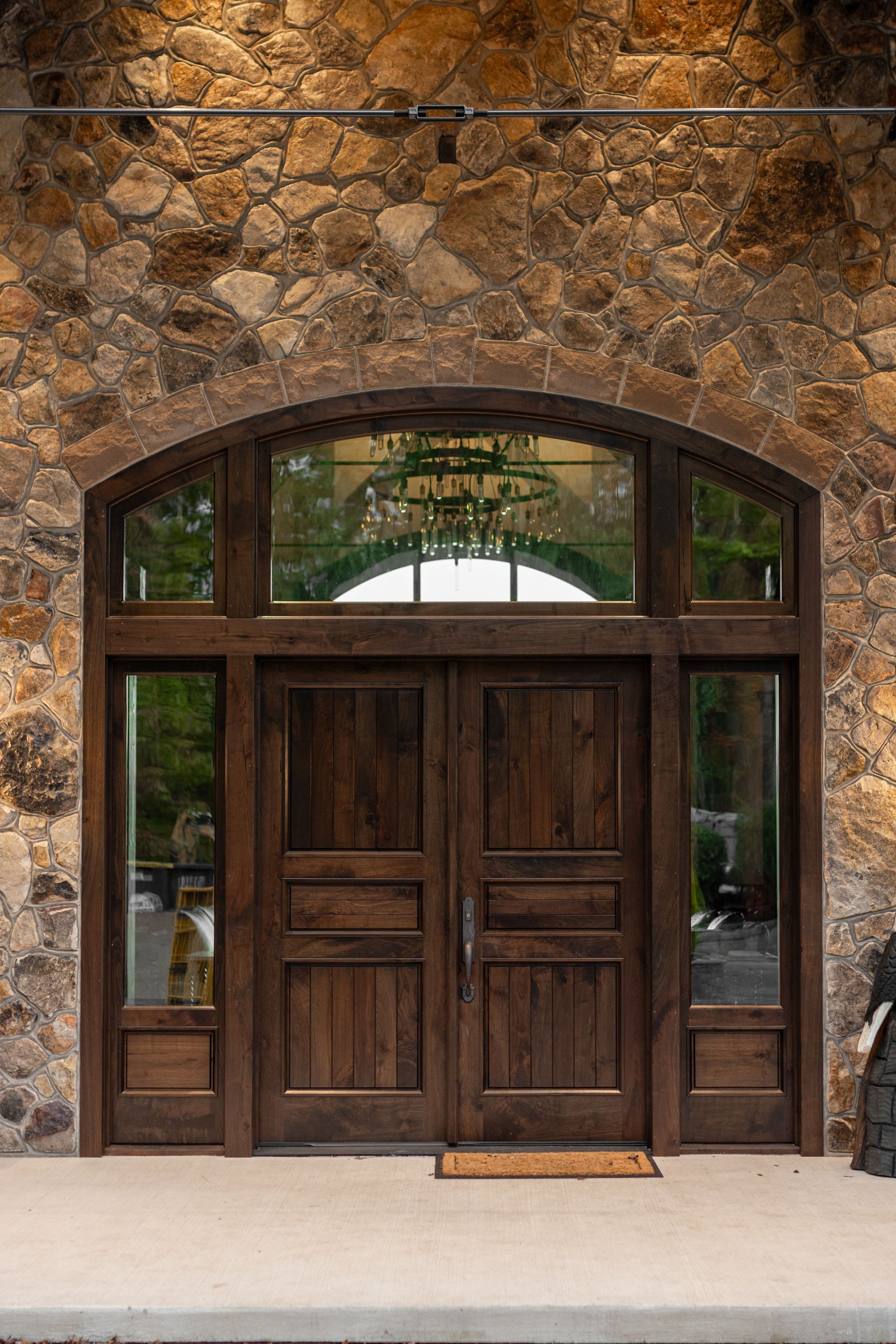 Brown wooden double doors with arched transom and sidelights in a stone wall.