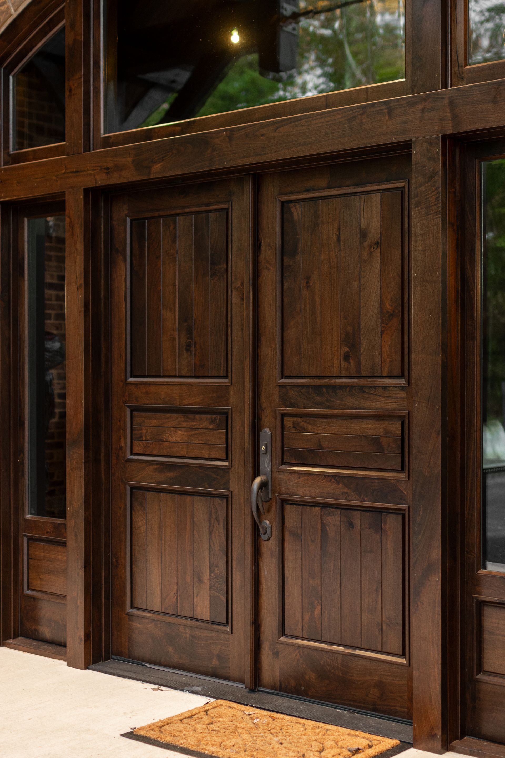 Dark stained wooden double doors with sidelights and a small doormat.