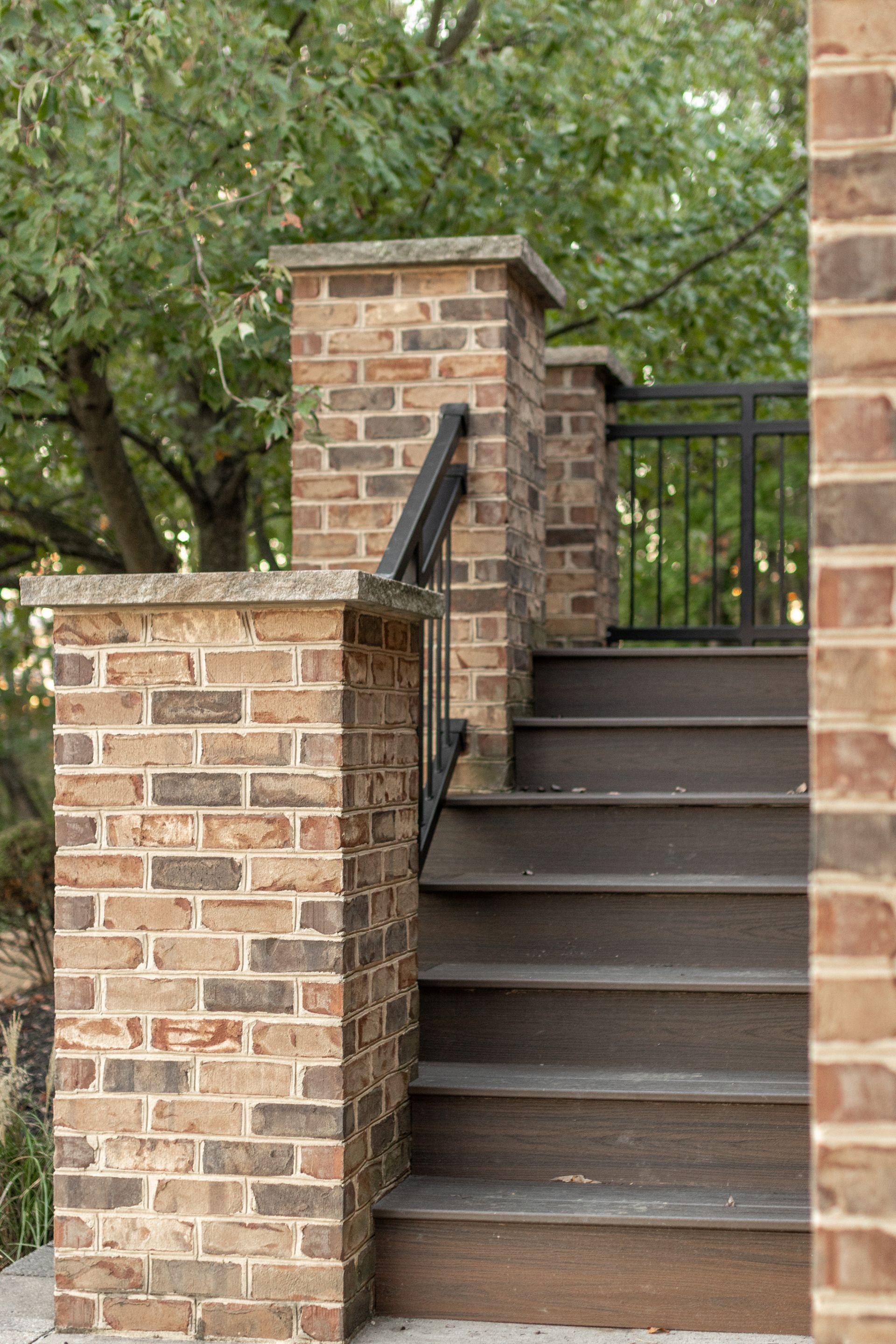 Brick staircase with black railing, leading to a gate, surrounded by brick pillars and trees.