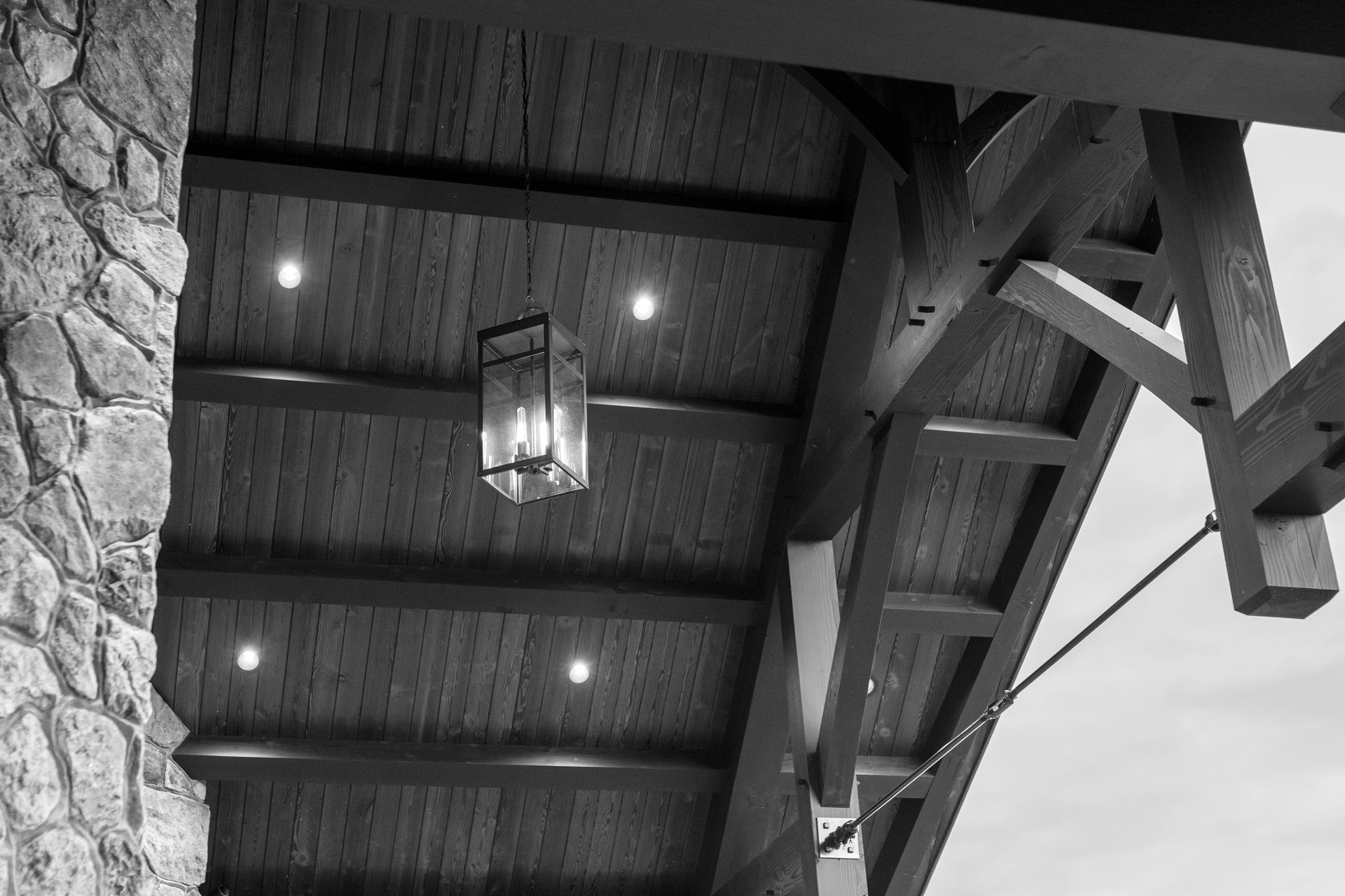 Black and white view of a wood beam ceiling with recessed lights and a hanging lantern. Stone column on left.