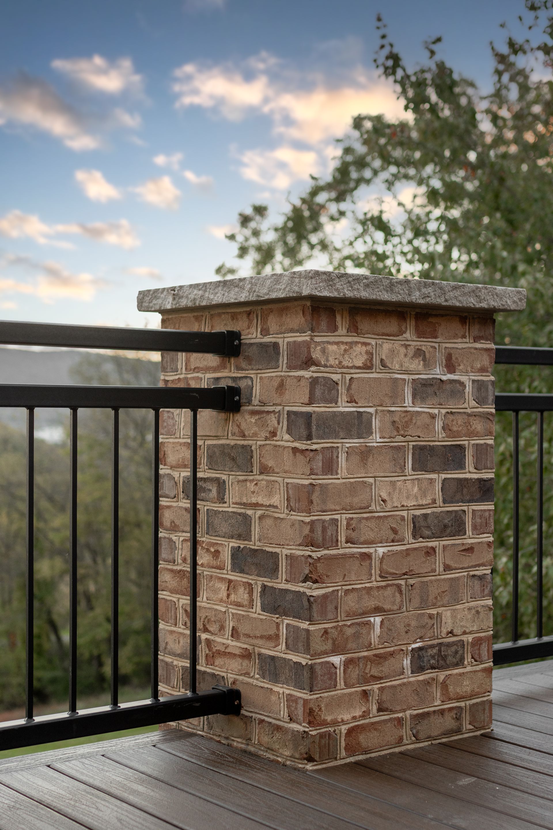 Brick pillar with black railing on a wooden deck, against a blurred sky and trees.