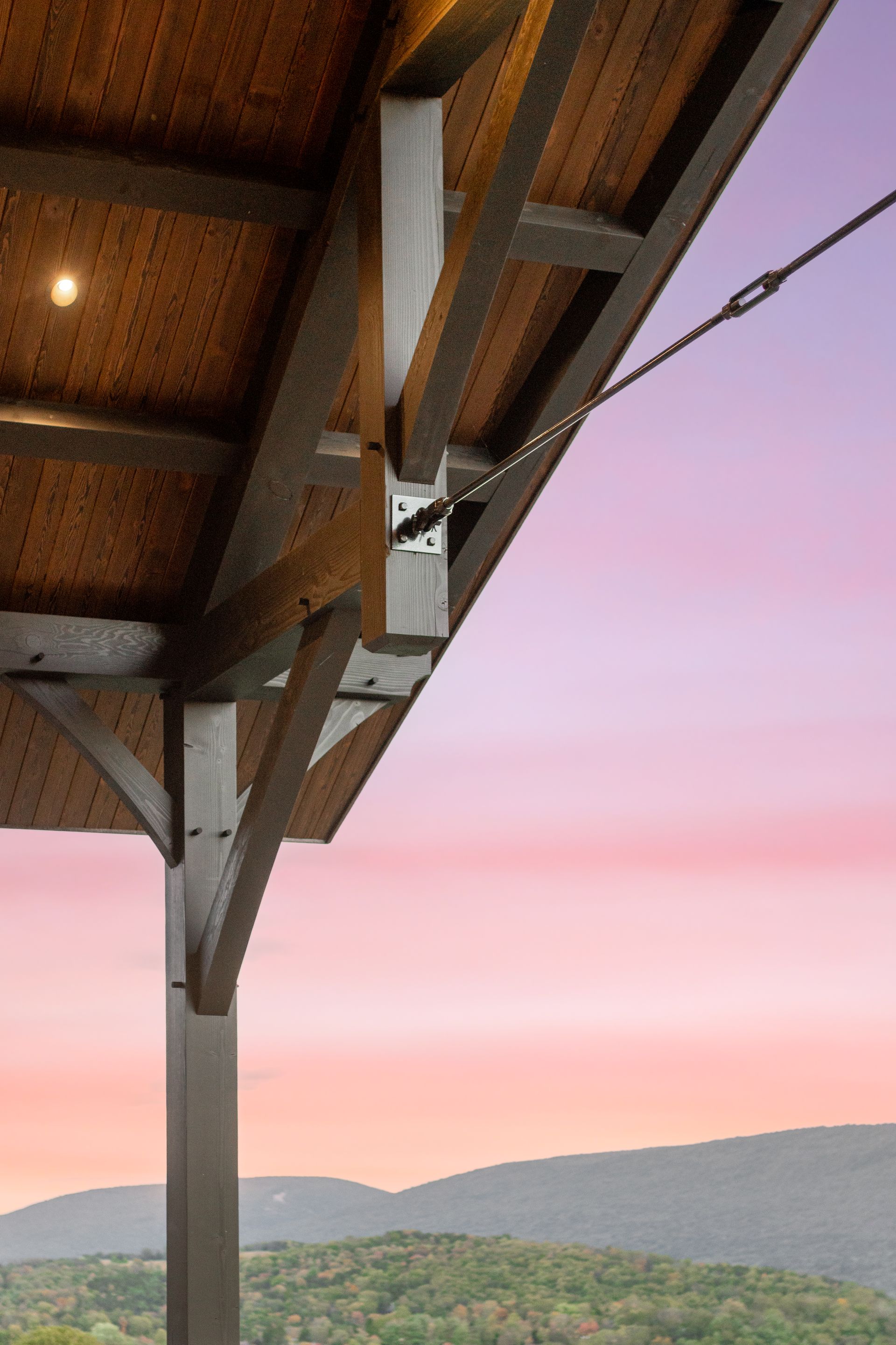 Wooden structure with exposed beams, support posts, and cables against a sunset sky over mountains.