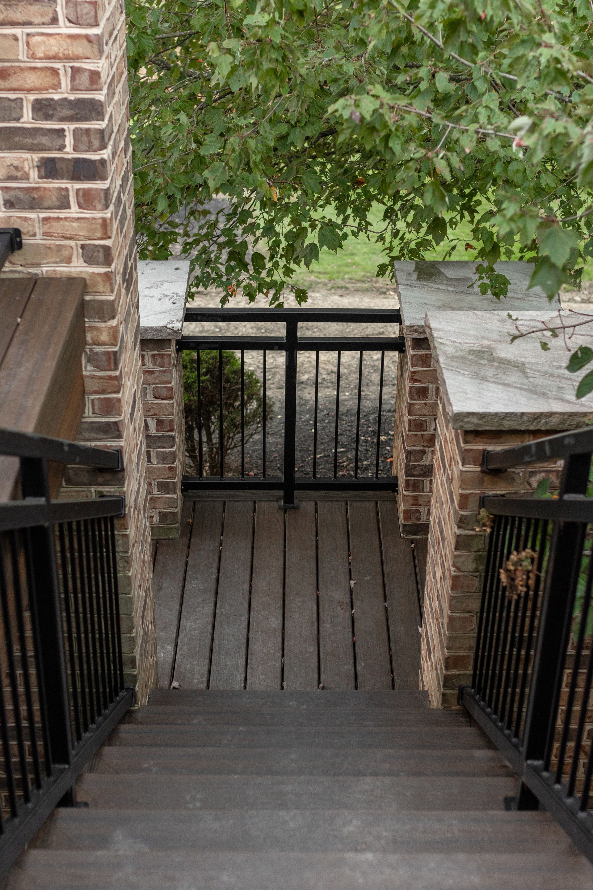 Stairs leading down to a wooden deck, flanked by brick columns and black railing, with a gated opening to a green space.