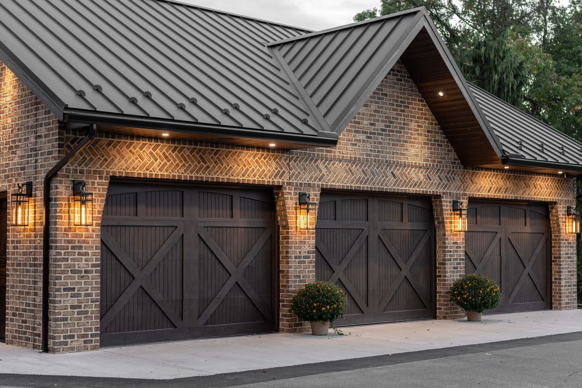 Brick garage with three dark wooden doors, metal roof, and warm exterior lights.