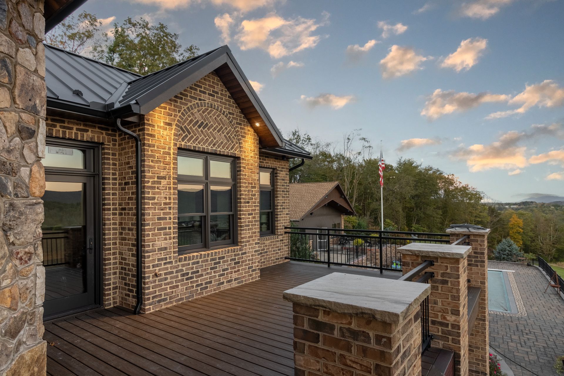 Brick house with a deck overlooking a landscape, pool and American flag under a sunset sky.