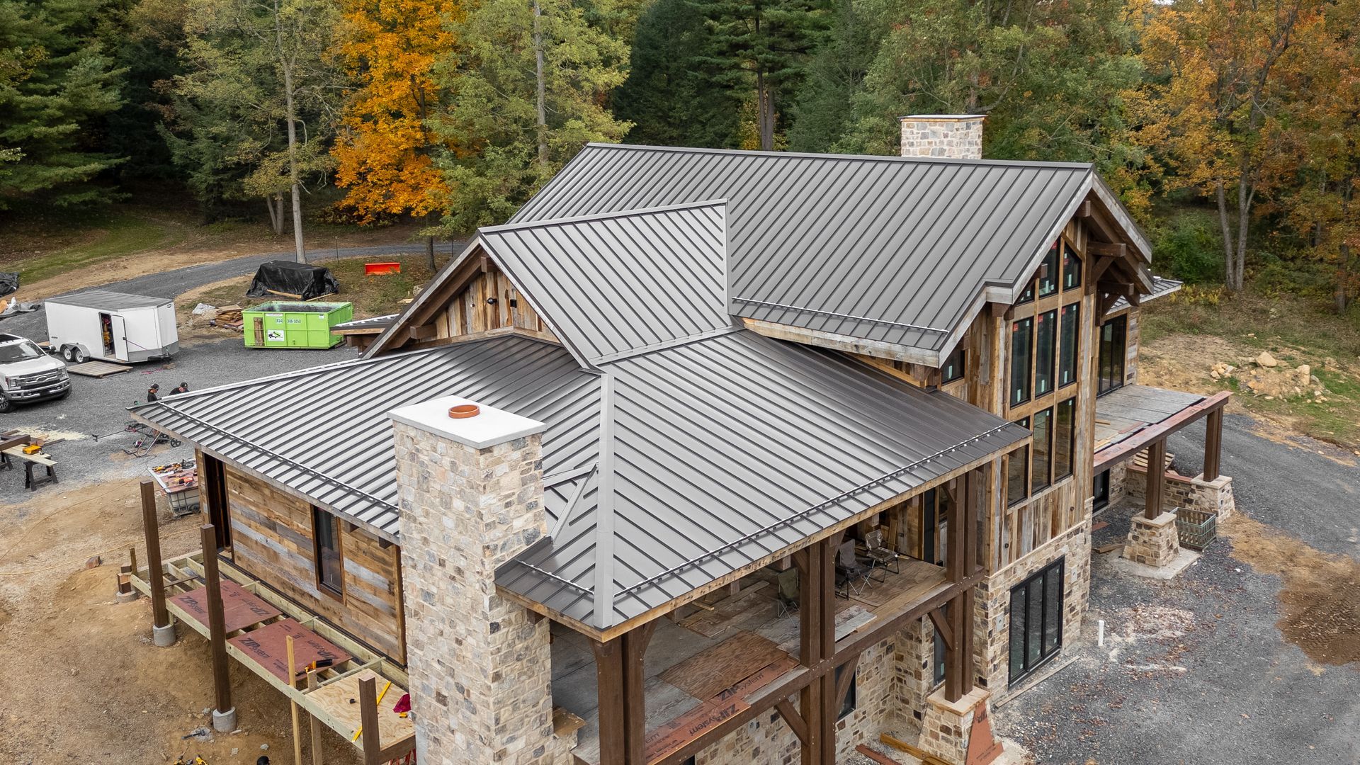 Rustic house with a metal roof under construction, with stone chimney and surrounding trees.