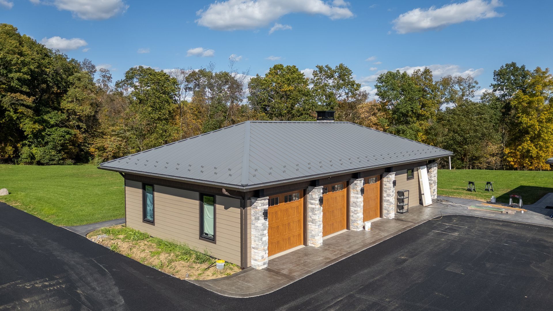 Tan, gray, and brown garage with stone accents and asphalt driveway, set against green lawn and trees.