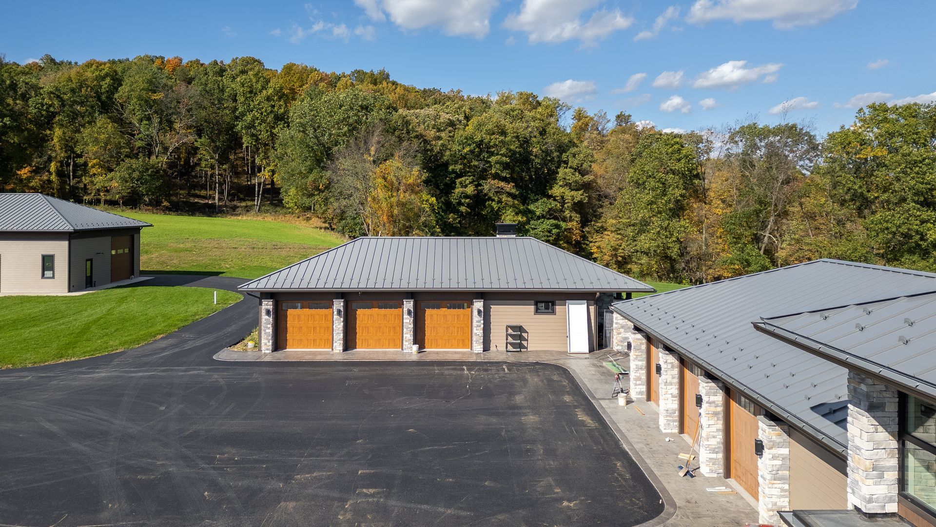 Three detached garages with asphalt driveway, tan siding, and brown garage doors; trees in the background.
