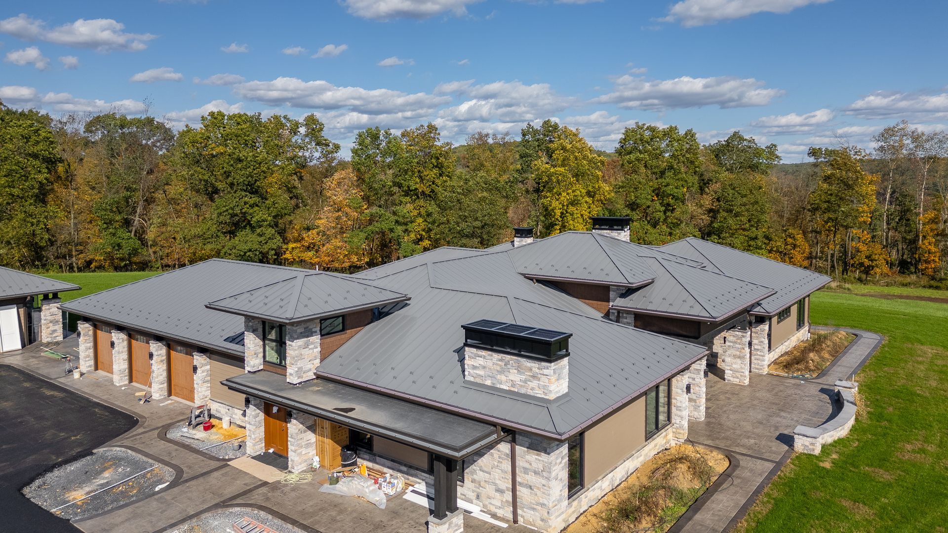 Modern single-story house with a gray roof, stone exterior, and a surrounding green lawn, near a wooded area.