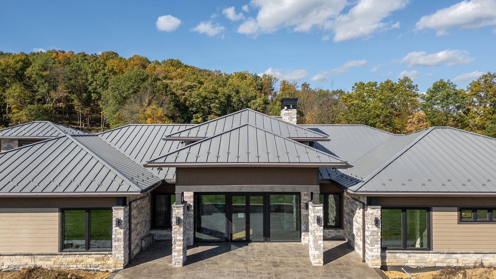 Modern home with gray roof and stone accents, set against a forested hillside and blue sky.