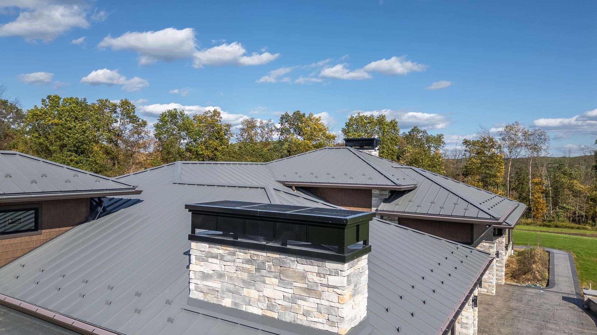 Grey metal roof of a house with stone chimneys, against a blue sky with scattered clouds.