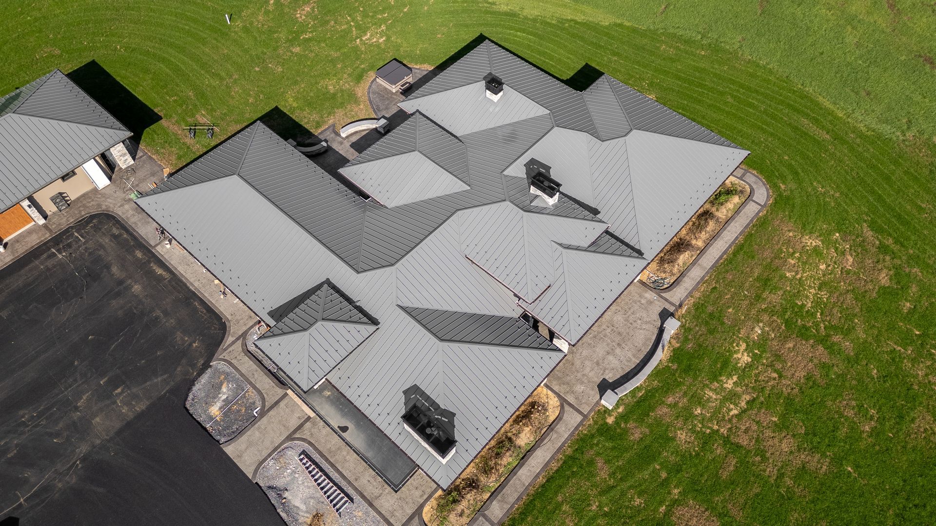 Aerial view of a modern house with a complex gray roof surrounded by green grass and a black driveway.