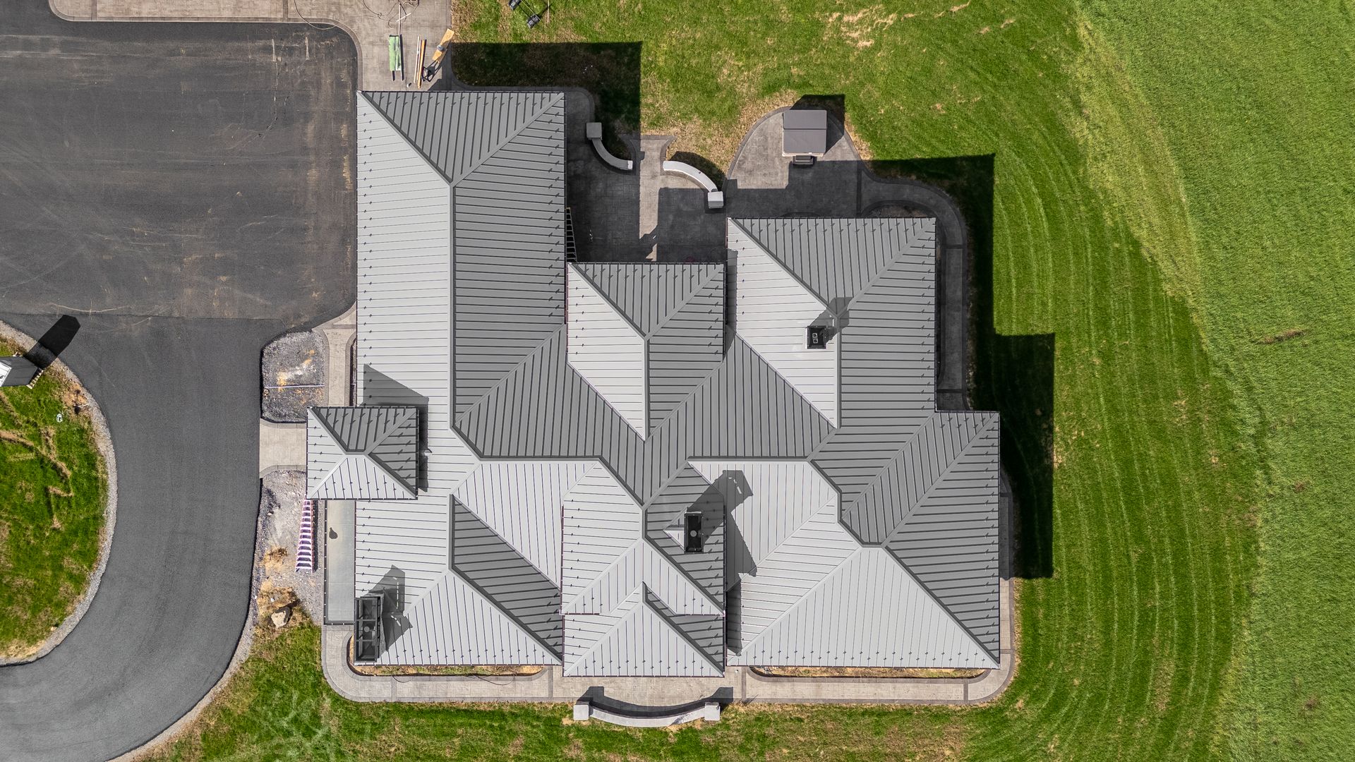 Aerial view of a house with a complex, multi-faceted grey roof next to a driveway and grassy field.