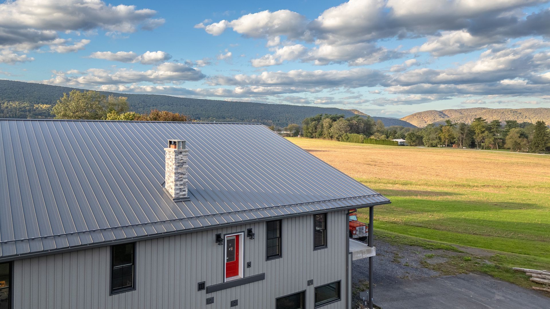 Two-story gray house with metal roof, red door, and chimney, set against a field and mountains under a cloudy sky.