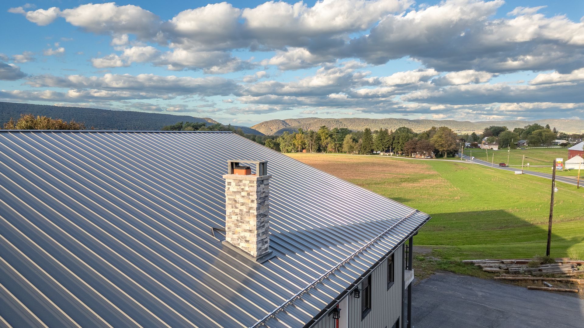 View of a metal roof with chimney, overlooking a green field and a mountain under a cloudy sky.