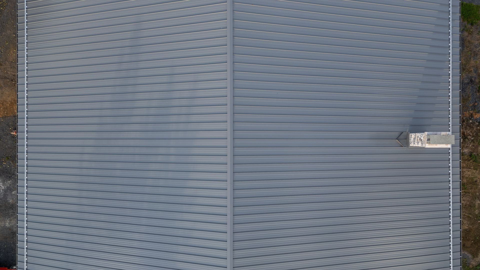 Overhead view of a large, gray, corrugated metal roof with a small building and chimney on the right.