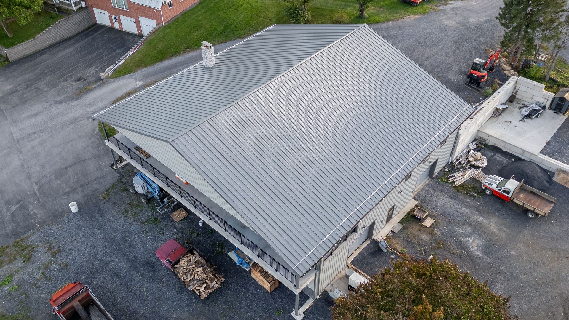 An aerial view of a two-story house with a gray corrugated metal roof, a gravel driveway, and several trucks.
