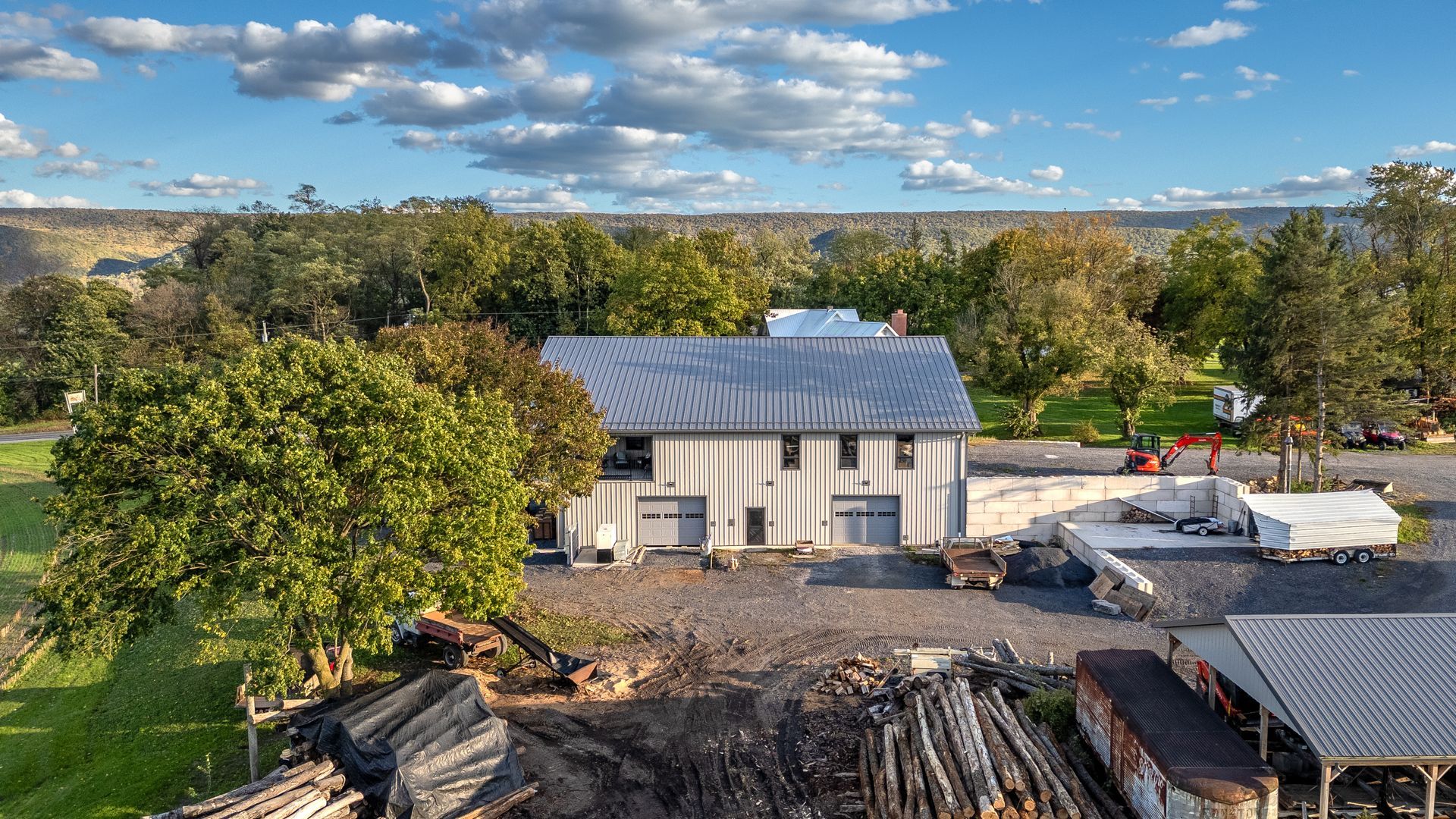 Two-story barn with gray metal roof and white siding, set on a lot with trees and construction equipment under cloudy sky.