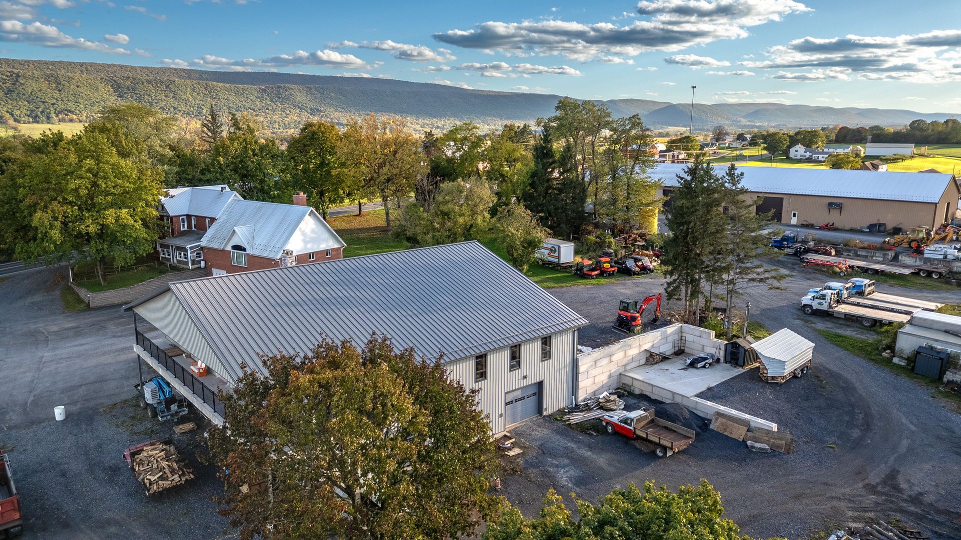 Aerial view of a large gray building with a metal roof, trees, vehicles, and mountains in the background under a blue sky.