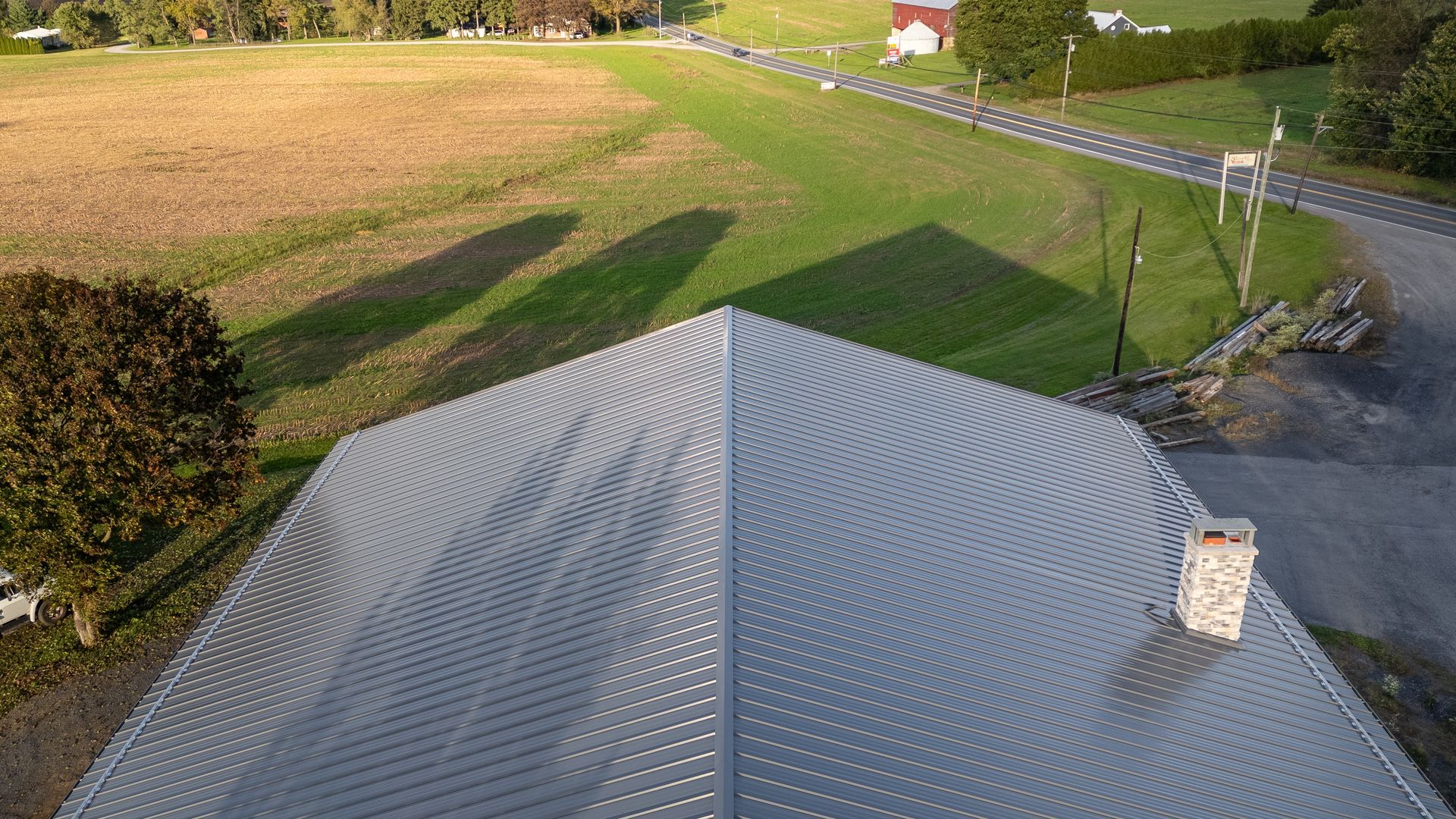 Gray corrugated metal roof with shadows, green field, road, and trees in the background.
