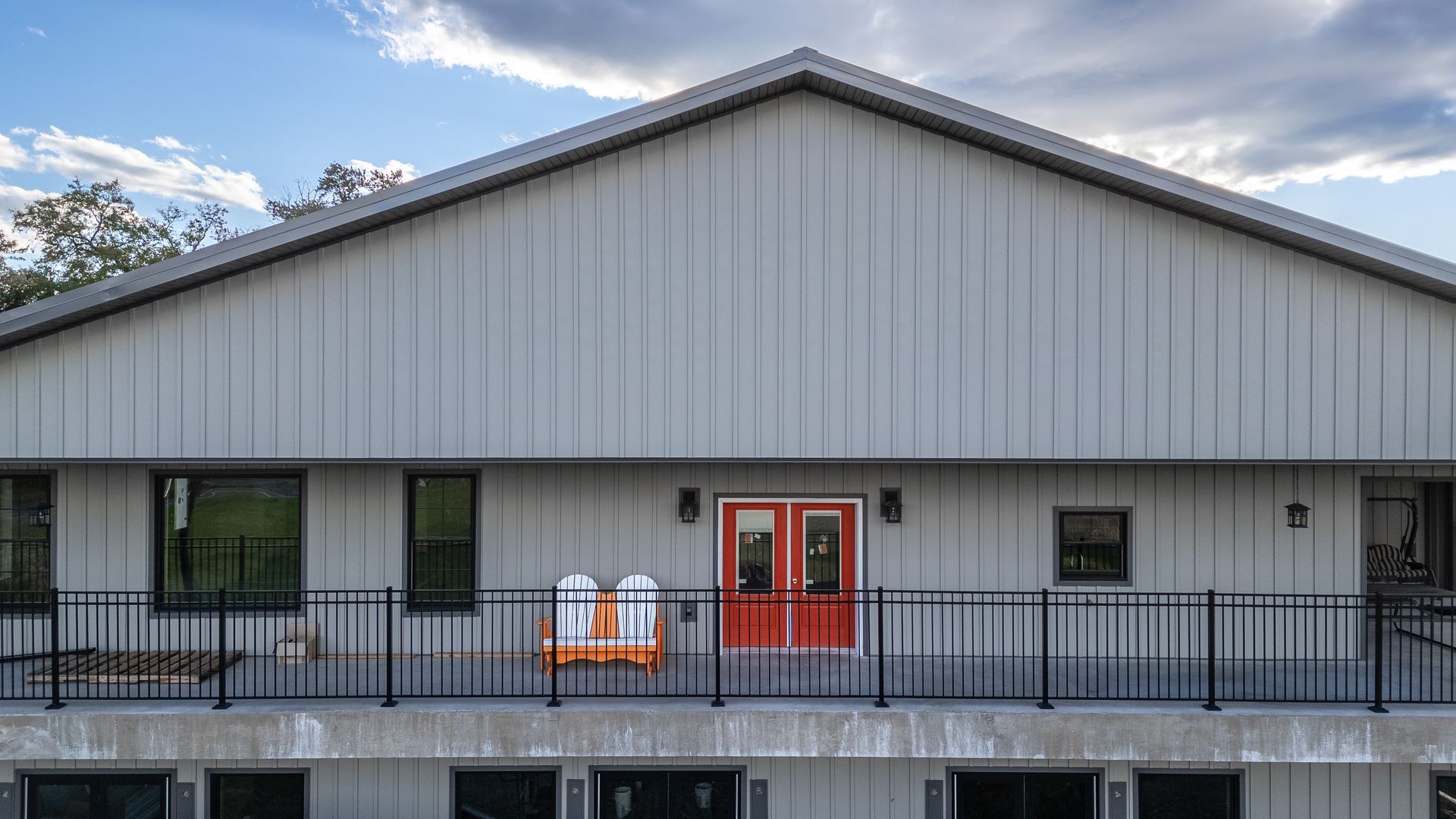 Two-story building with gray siding, balcony, black railings, orange double doors, and white chairs.