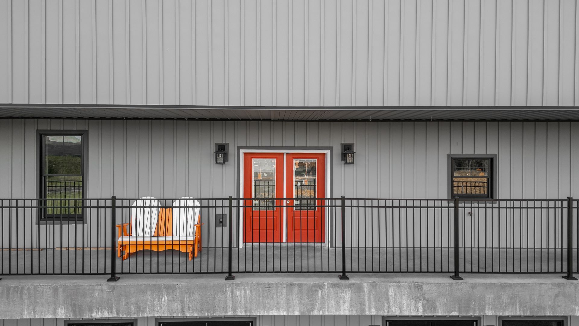 Exterior of a building with gray siding, red double doors, a black railing, and an orange bench.