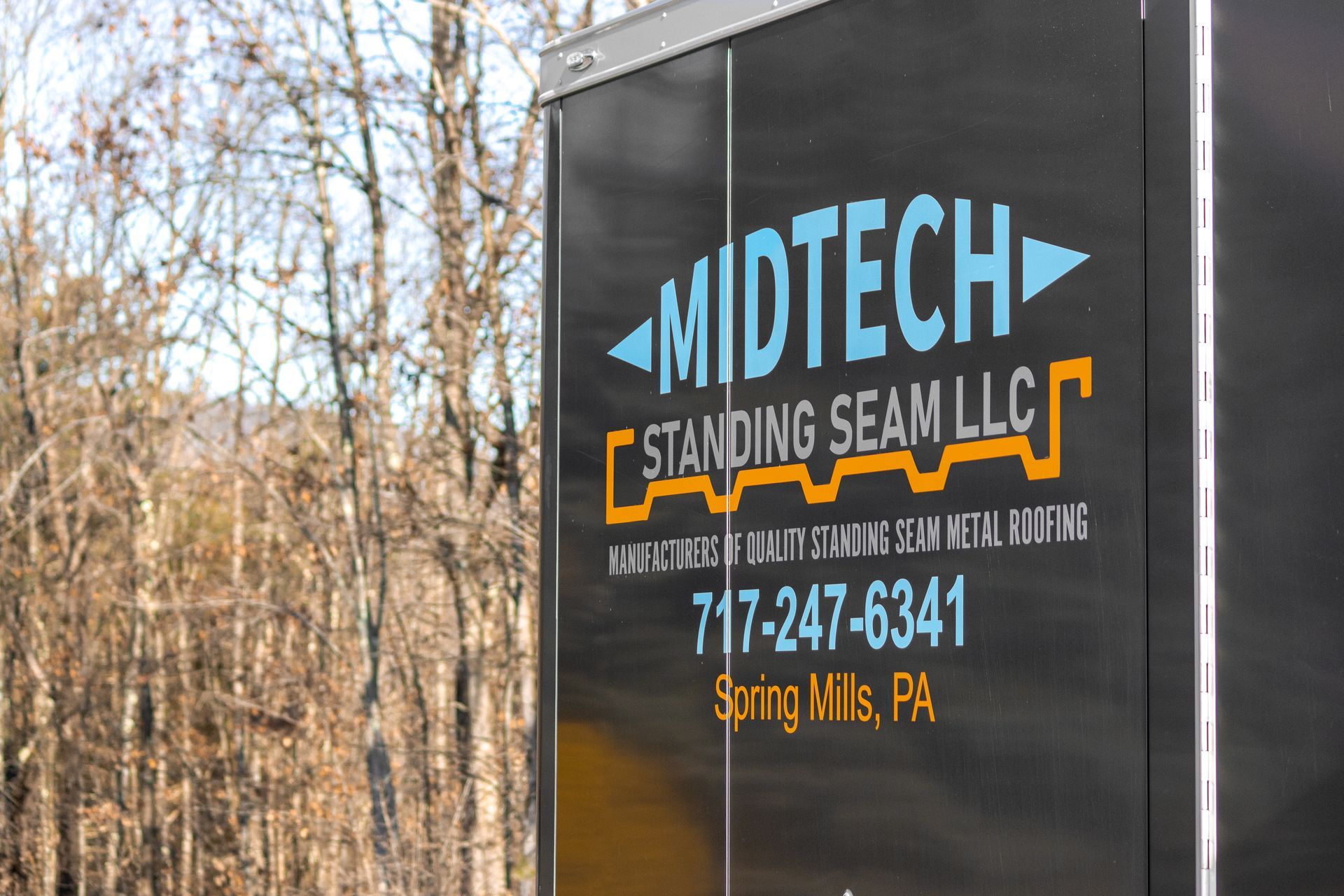 Black trailer with Midtech Standing Seam LLC logo and contact info in blue, orange, and white; trees in background.
