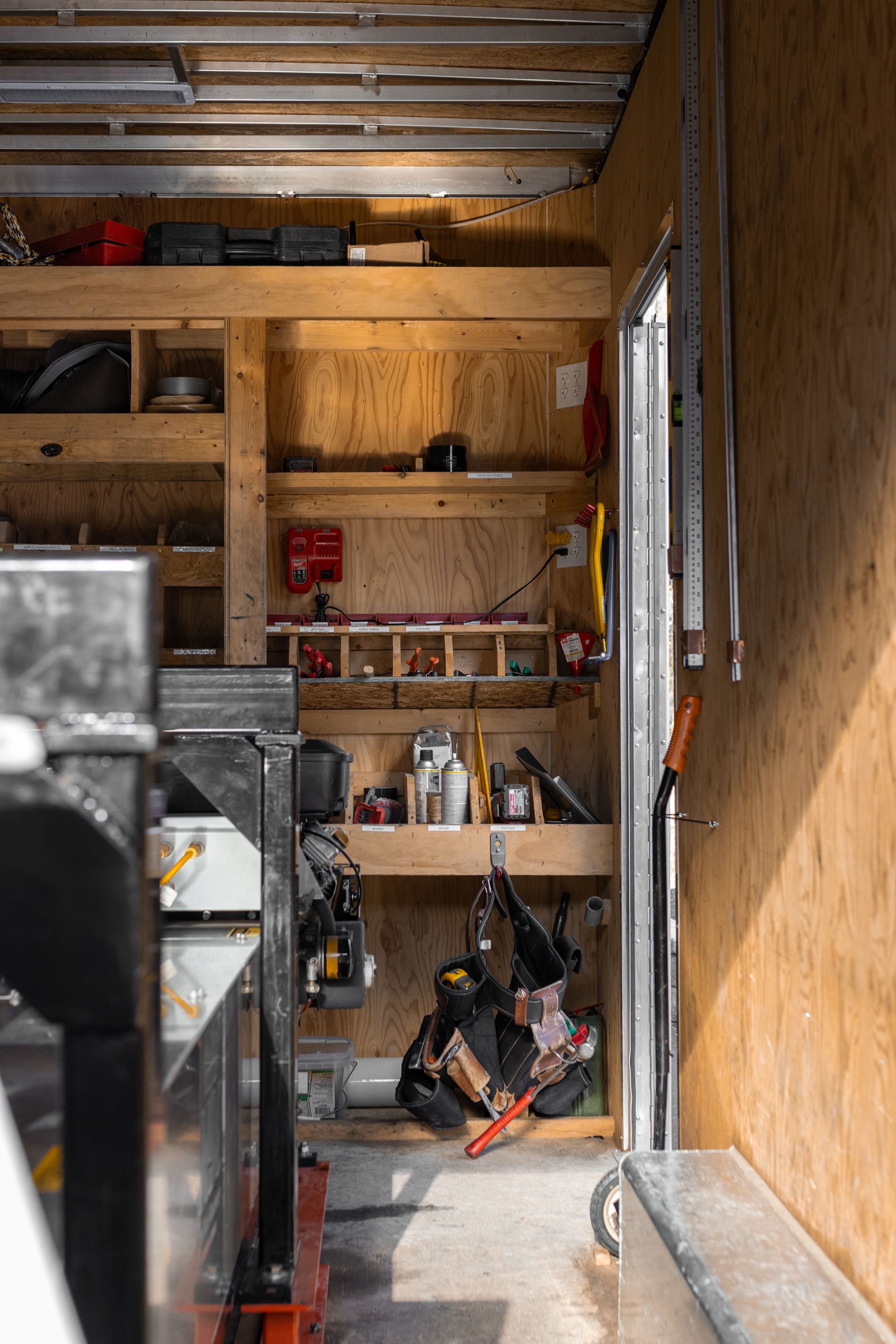 Interior of a workshop with wooden shelves. A person works below hanging tools. Sunlight shines through the open doorway.