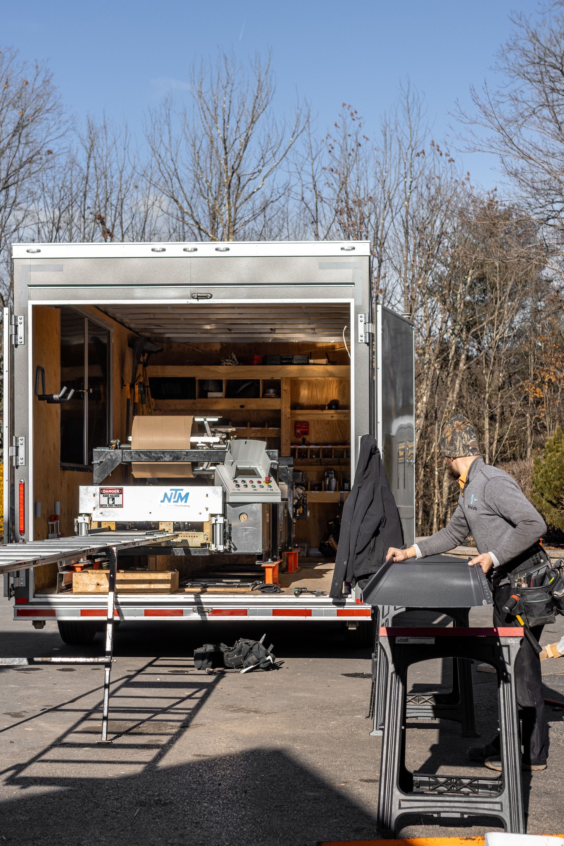 Man working at a trailer workshop. He's standing near a saw table with the open door revealing tools and shelves inside.