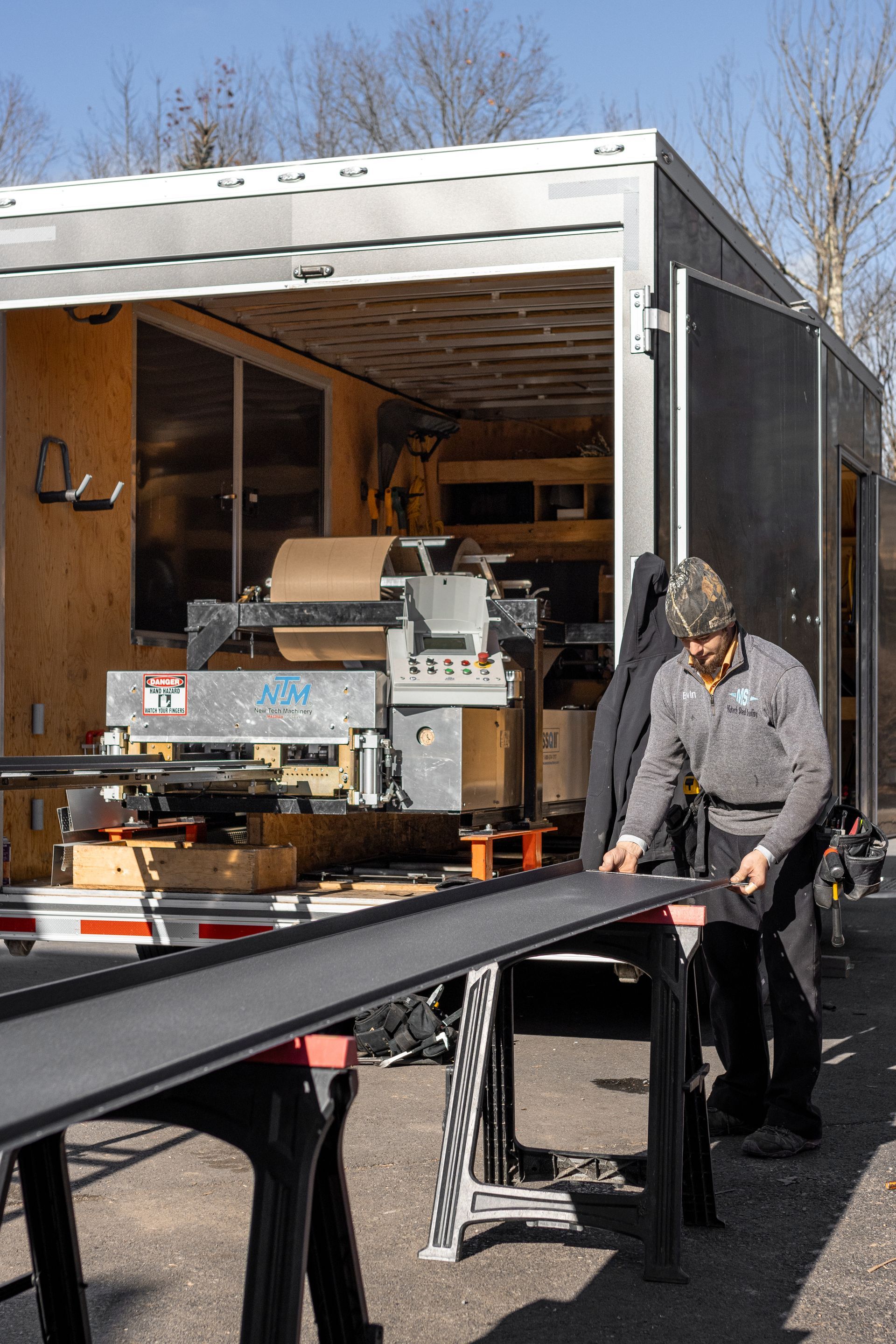 Man working with long black material on sawhorses outside a trailer.