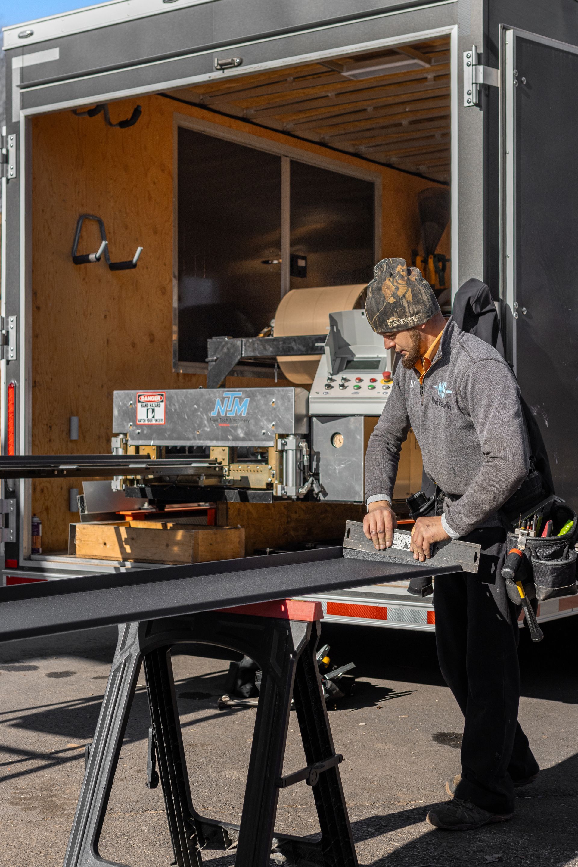 Man working on metal, likely for a window, outside a trailer. He wears work clothes and a tool belt.
