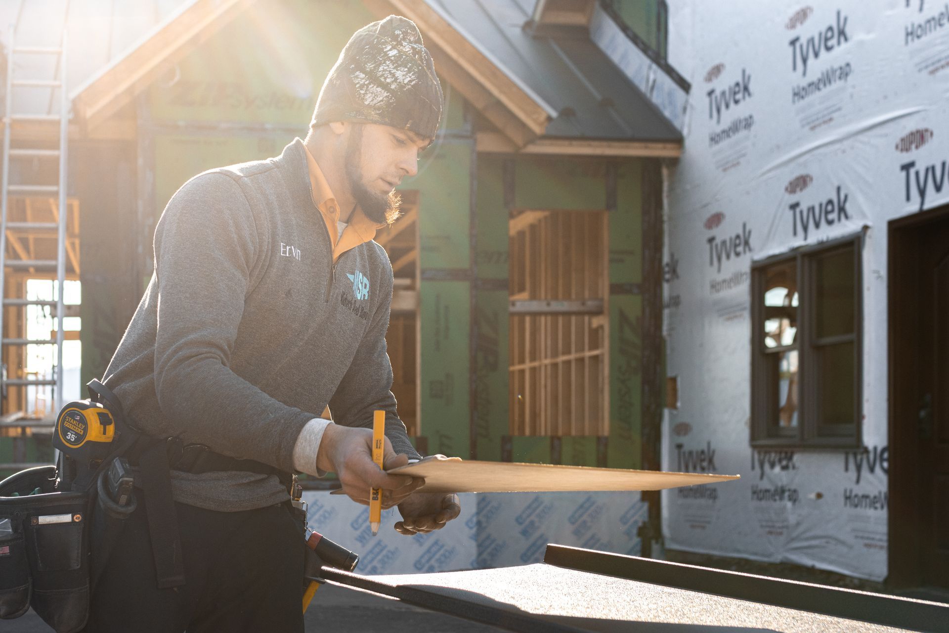 Construction worker inspecting wood at a building site; wearing a beanie, gray shirt, and tool belt.
