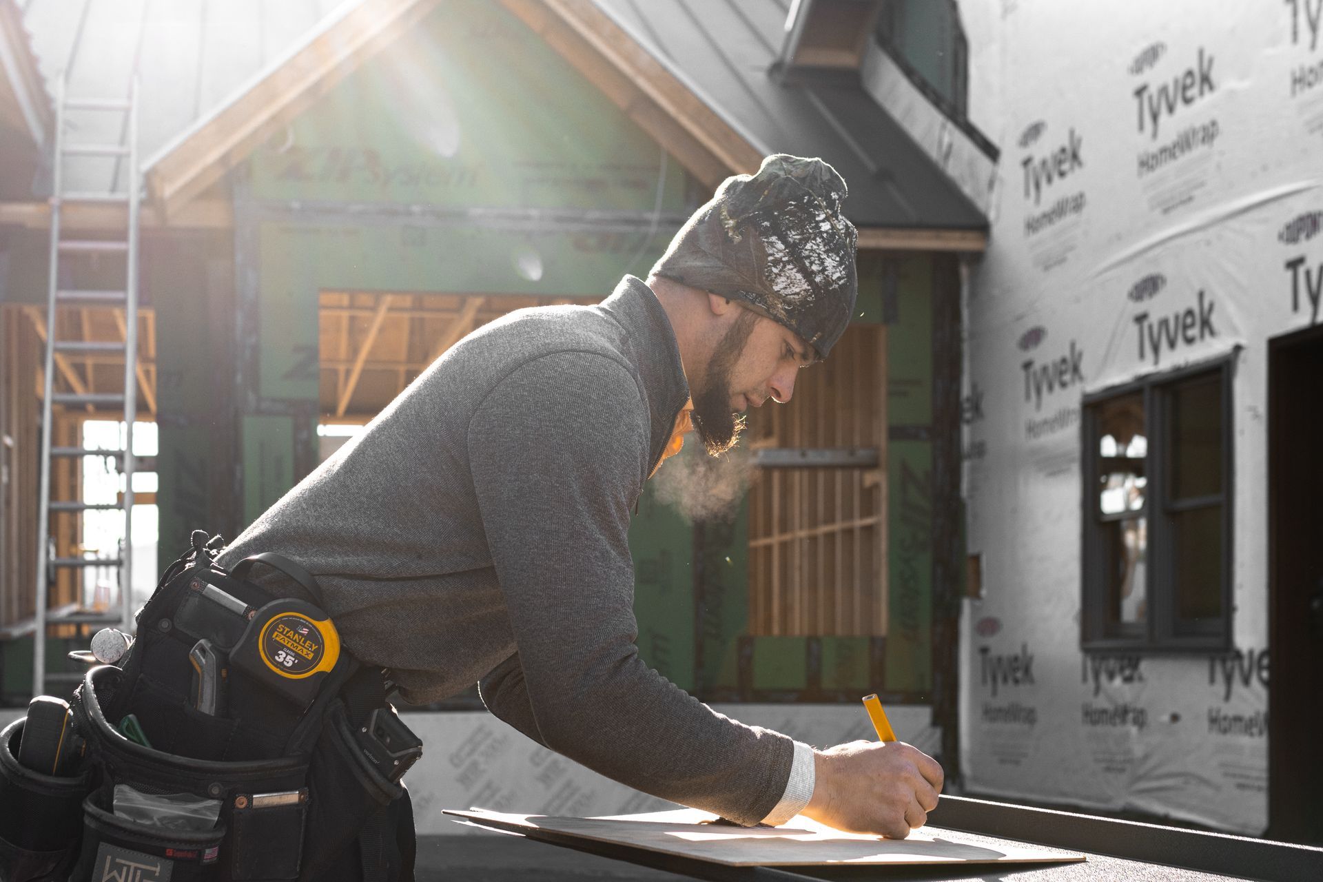 Construction worker in beanie, marking a plan, with a partially built house in the background.