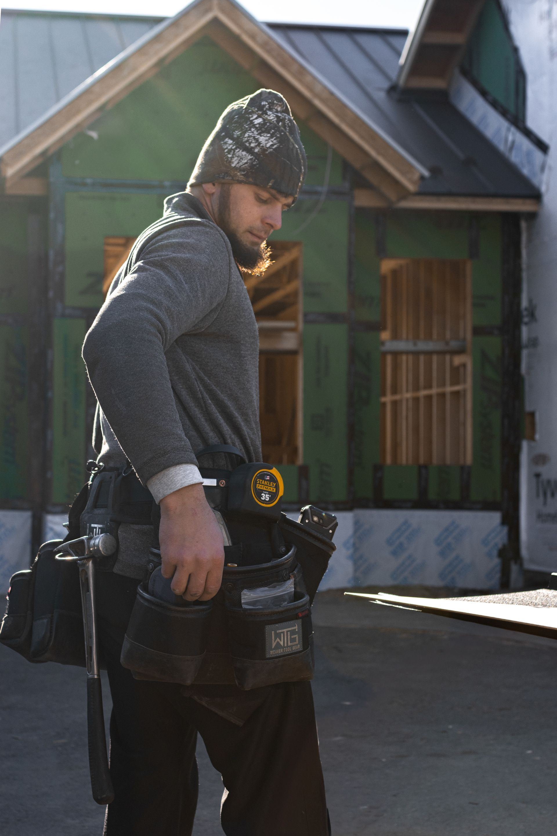 Construction worker with tool belt, looking down, in front of a house under construction.