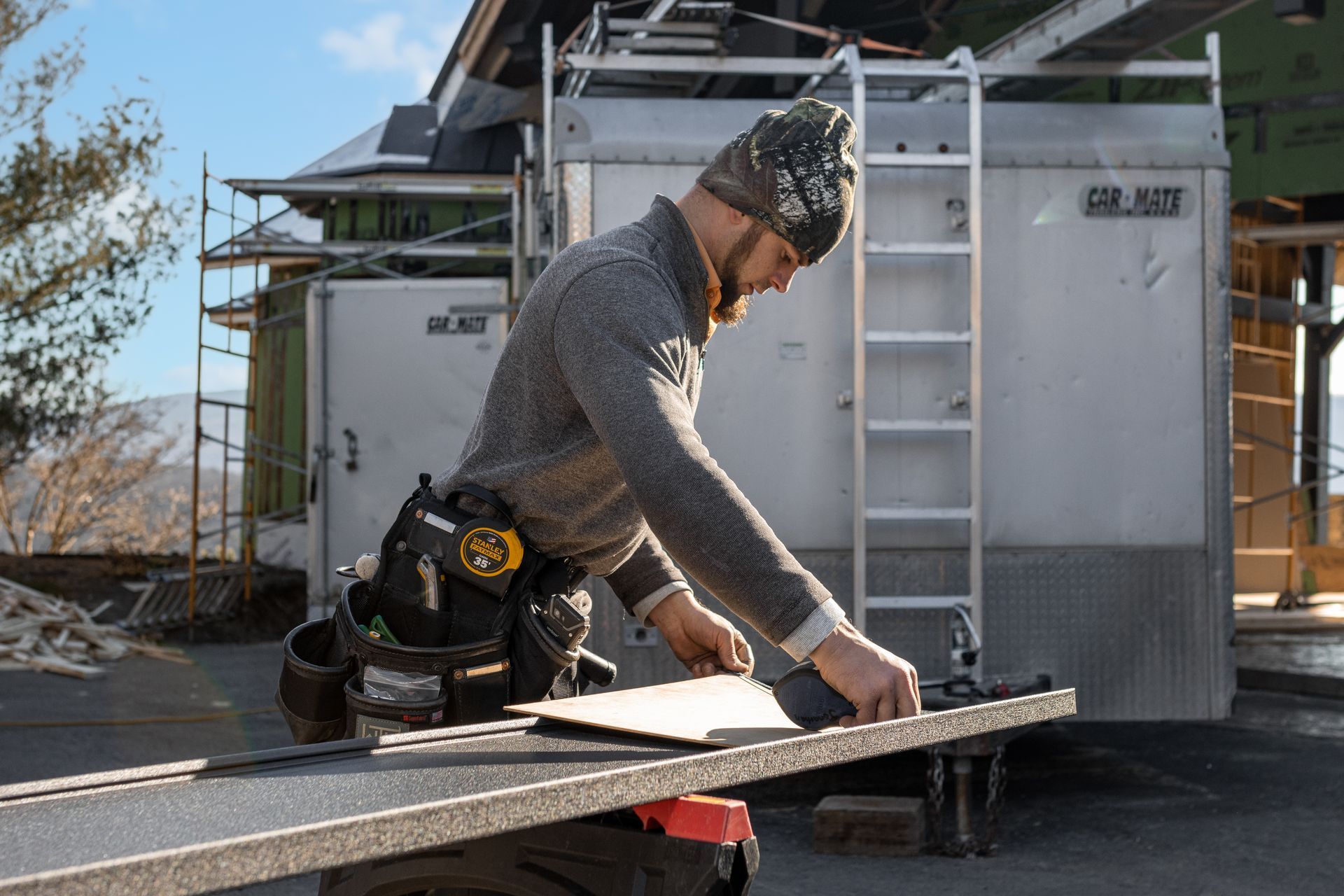 Carpenter in beanie measuring a board outdoors, tool belt visible. Trailer and building in background.