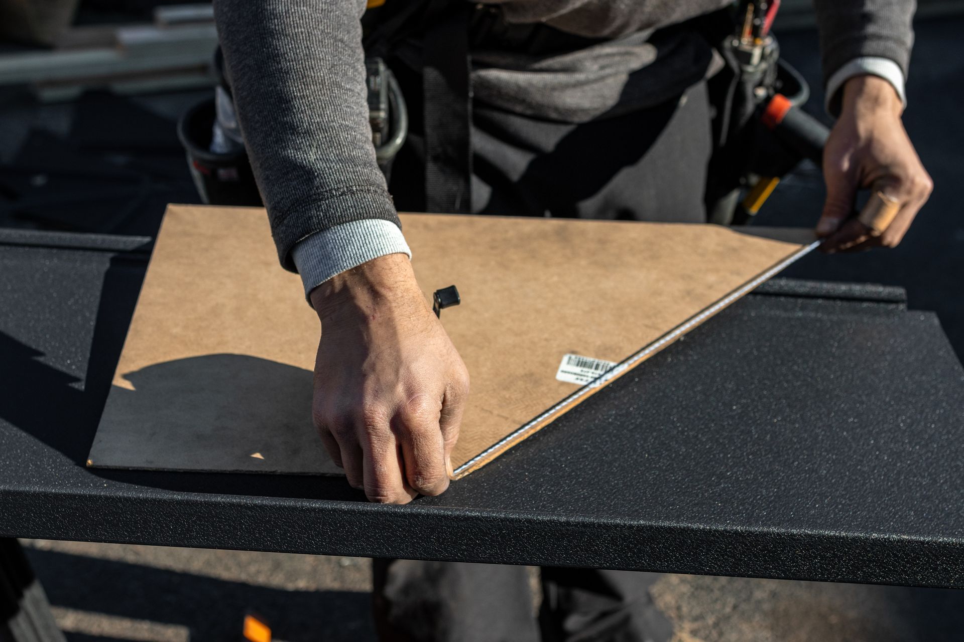 Person cutting cardboard on a black surface; wearing gray sweater, work pants, and tool belt.
