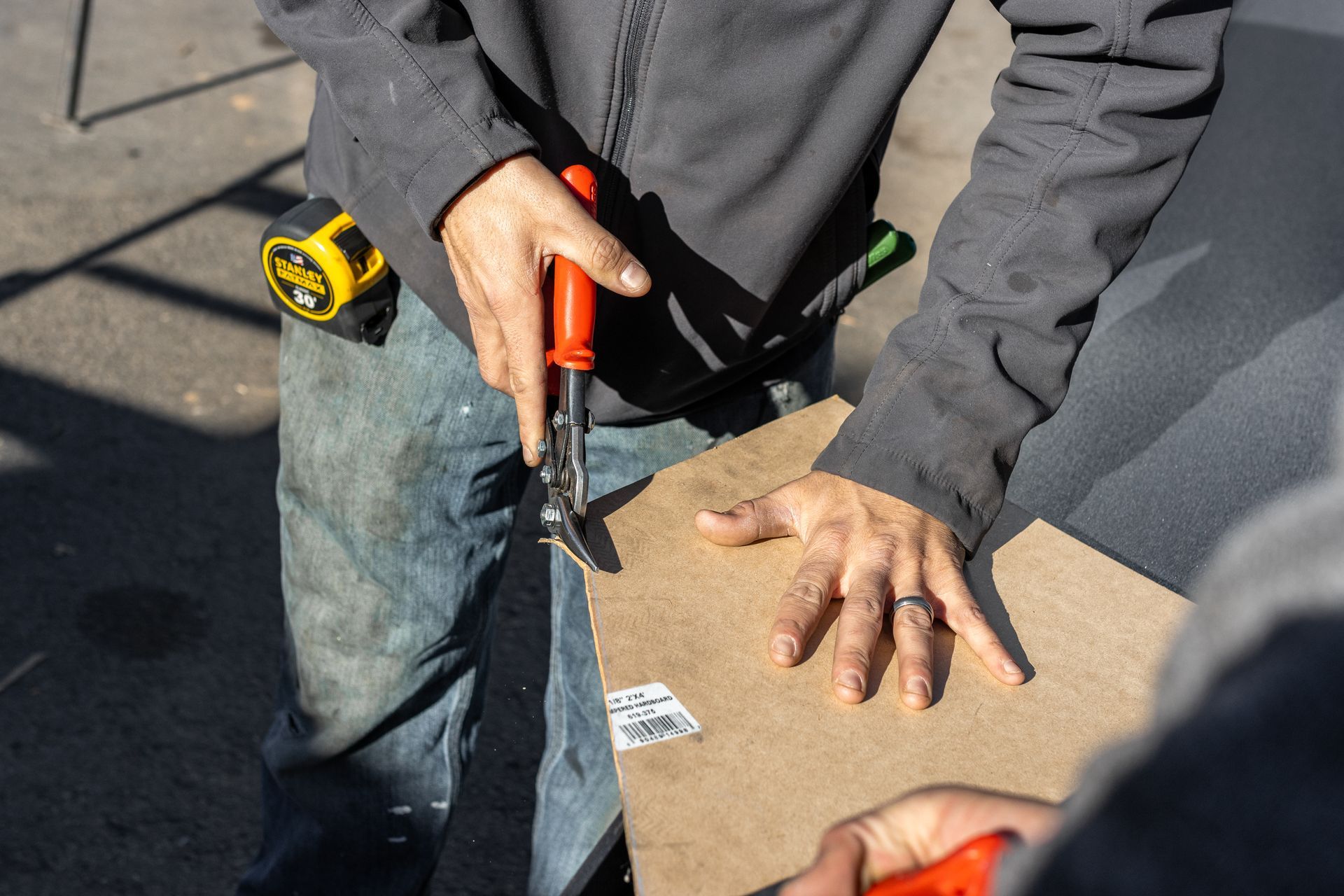 Person cutting a piece of cardboard with tin snips outdoors, holding a measuring tape.