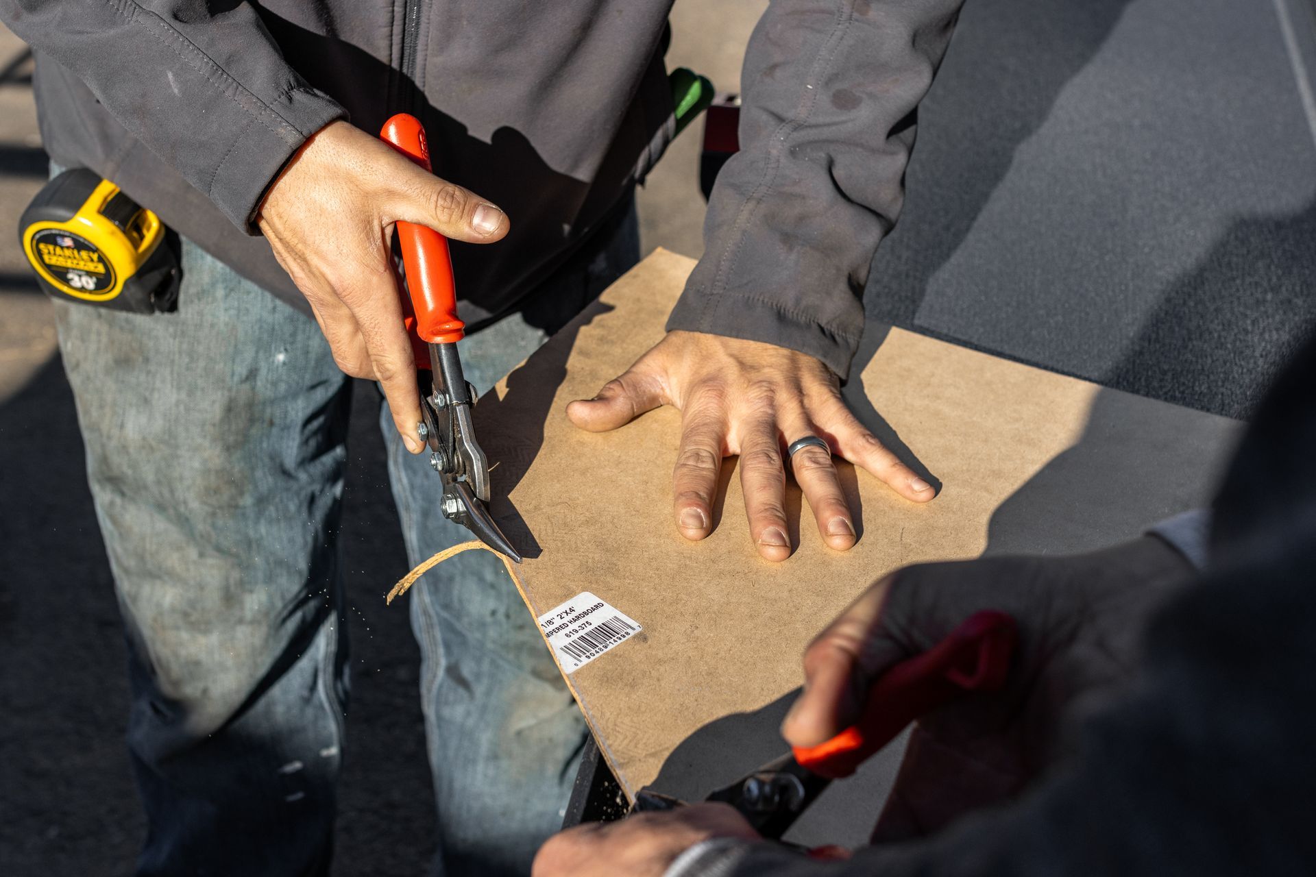 Two people cutting cardboard with red-handled snips; one wears a ring and jeans; outdoors.