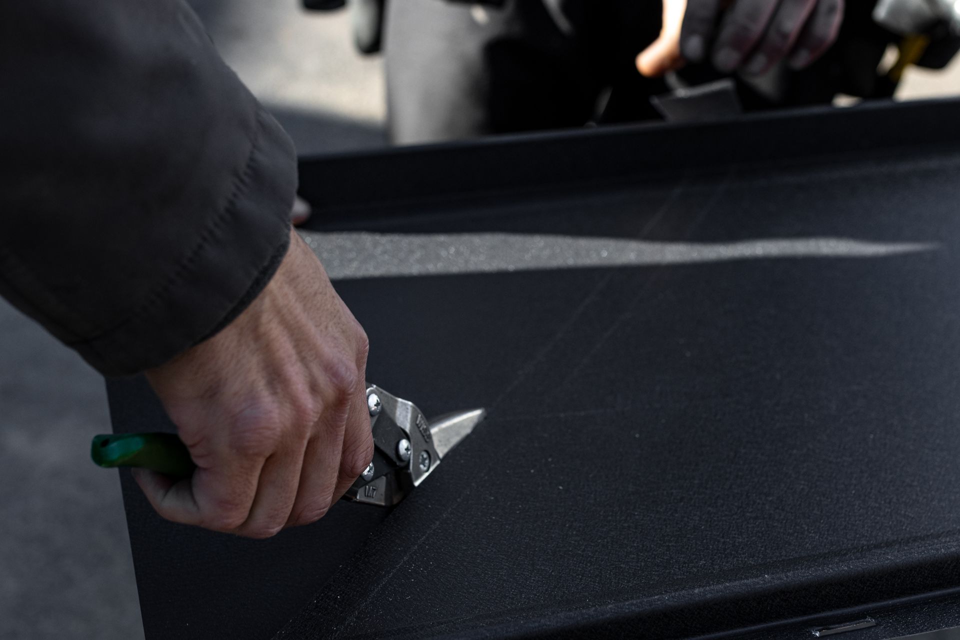 A person using metal shears to cut a black, textured sheet, outdoors.