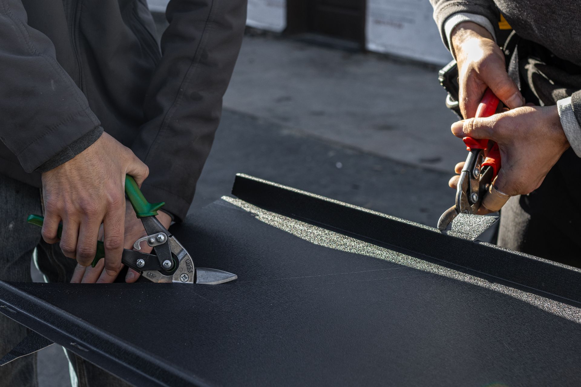 Two people using metal snips to cut a black metal sheet outdoors.