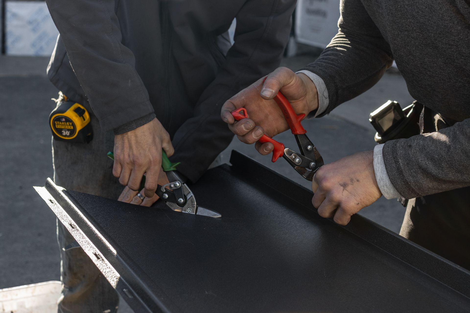 Two people cutting black metal with shears outdoors. One uses green, the other red tools.