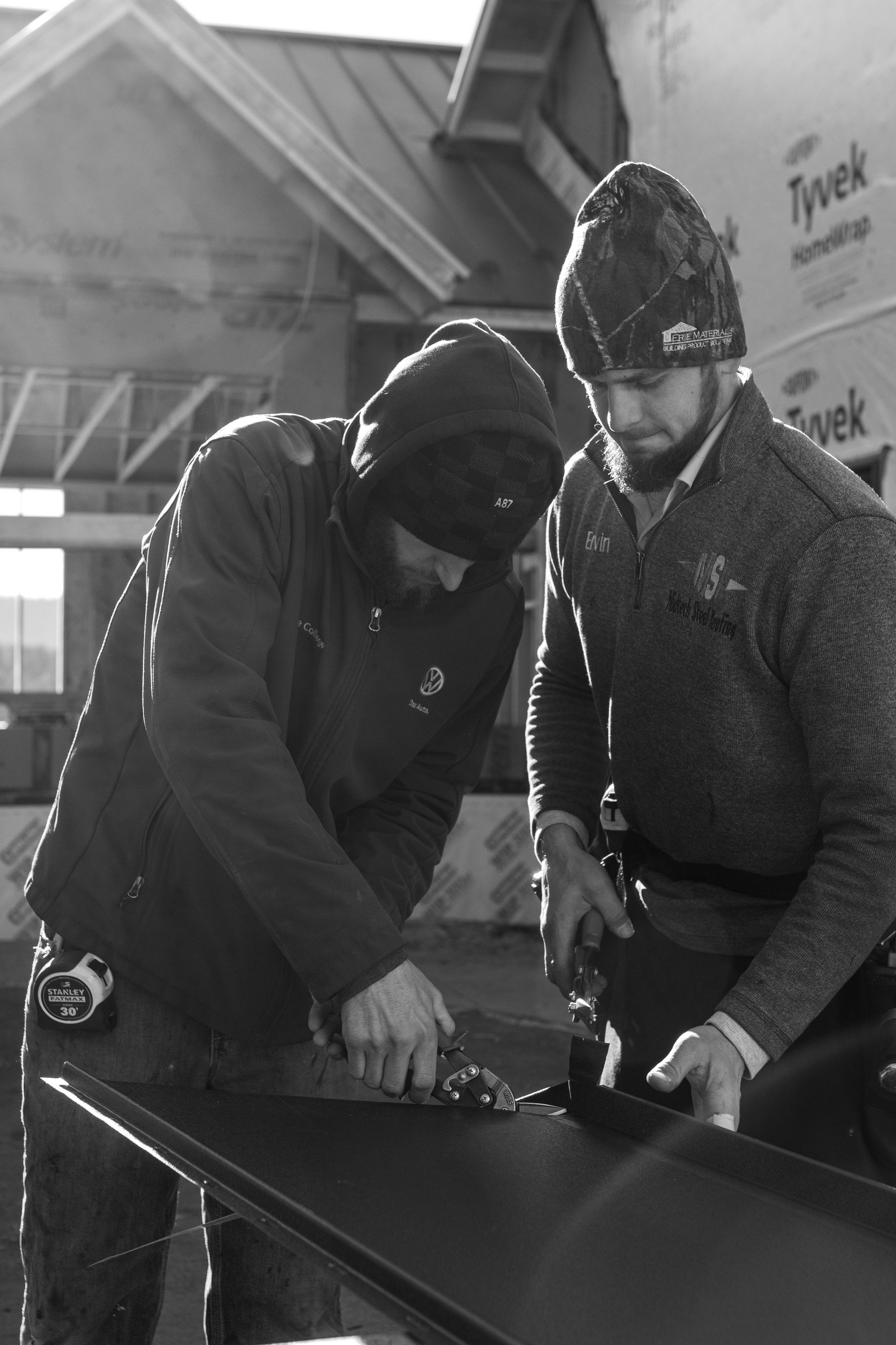 Two construction workers cutting metal roofing material outdoors.