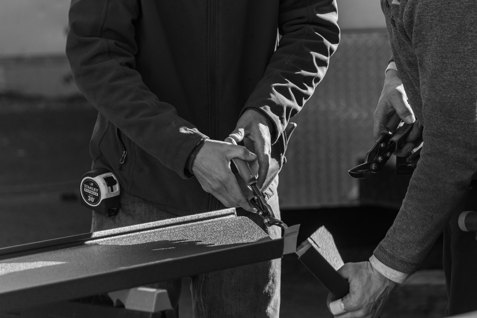 Two people working on a project outdoors. One uses pliers, the other holds a hammer.
