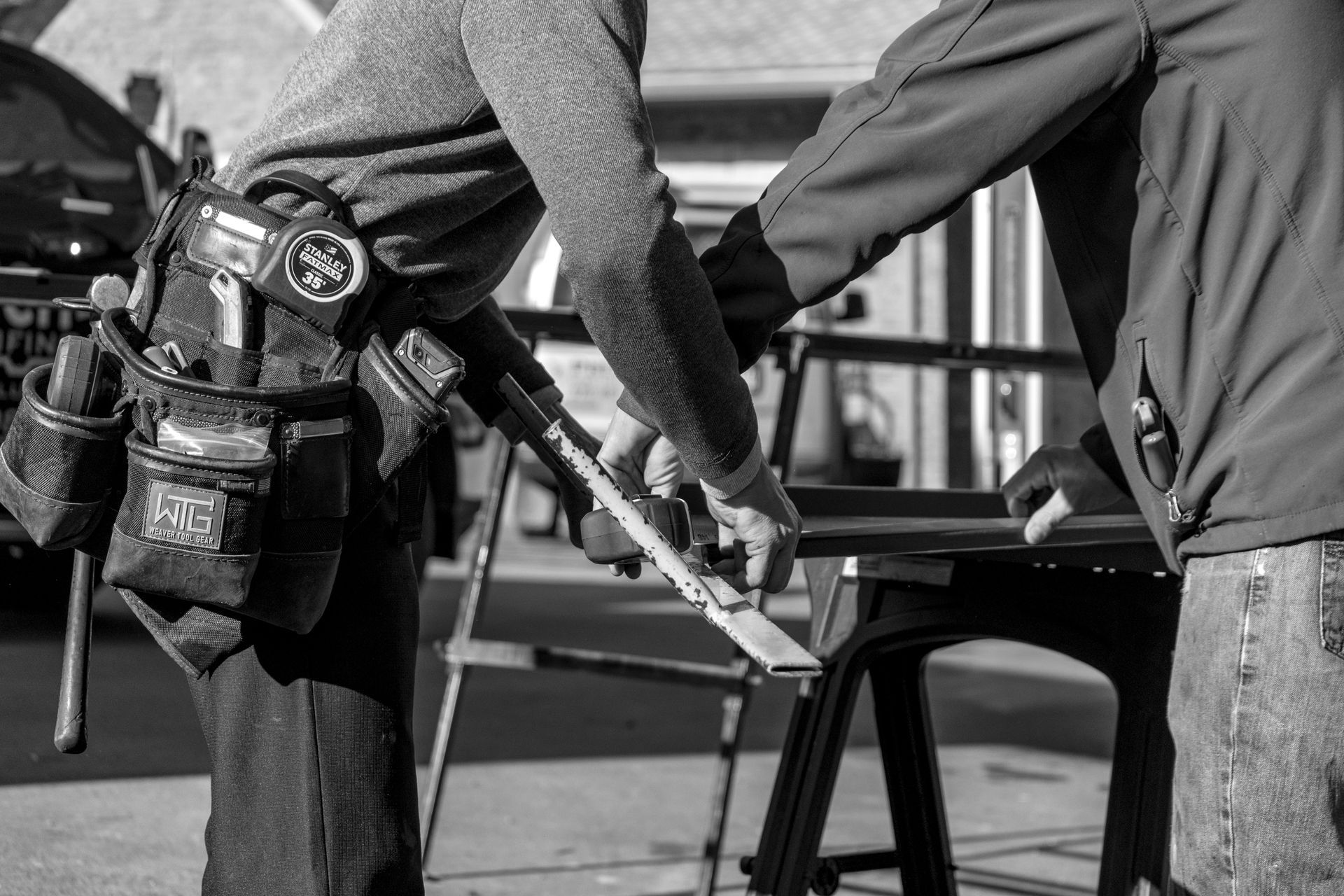 Two people working on wood outdoors. One cuts wood with a saw while another assists. Black and white.